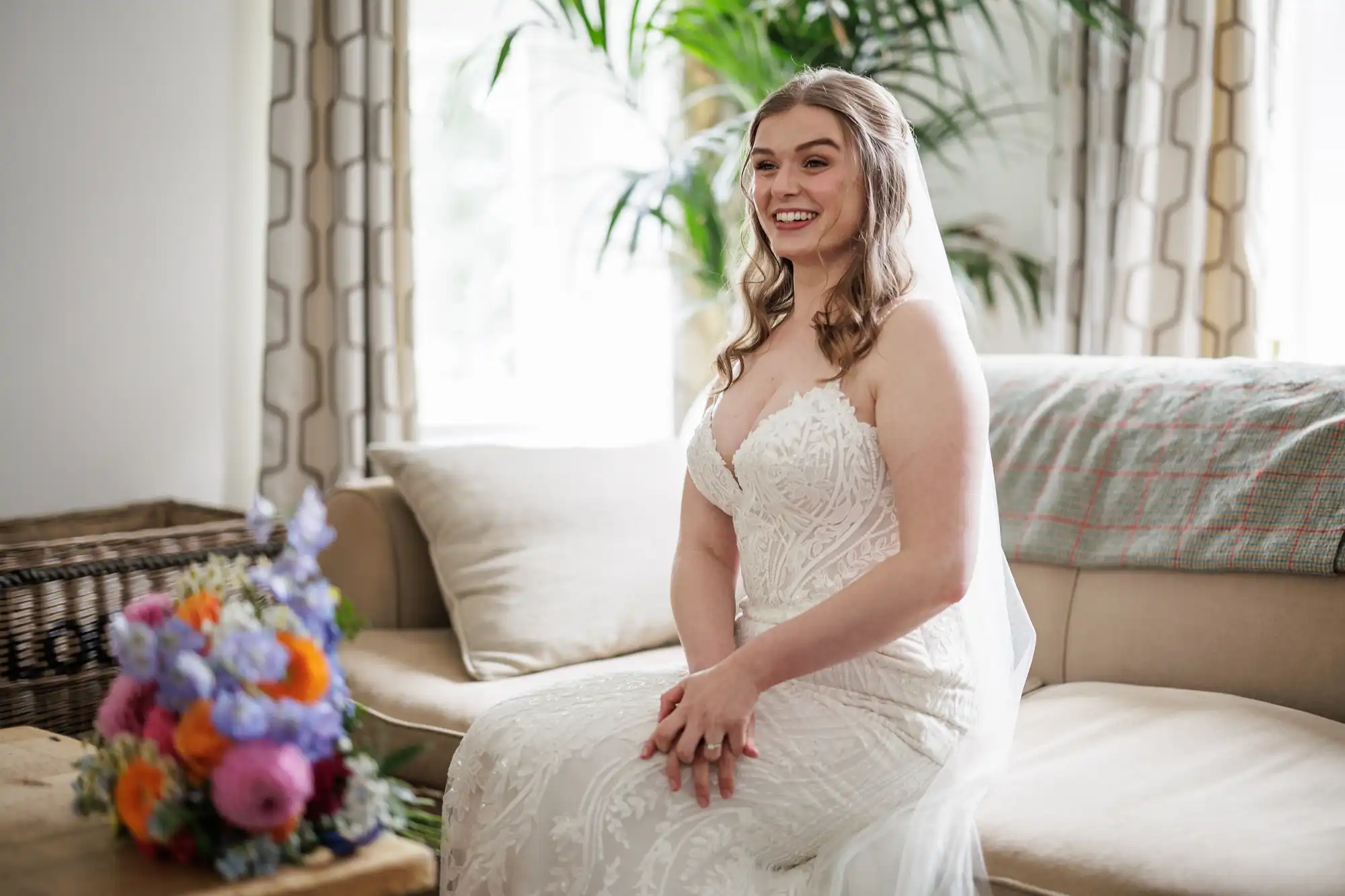 A bride in a white lace wedding dress sits smiling on a beige sofa, hands clasped on her lap. A colourful bouquet of flowers rests on a wooden table nearby. Sunlight filters through windows and plants in the bright, cosy room.