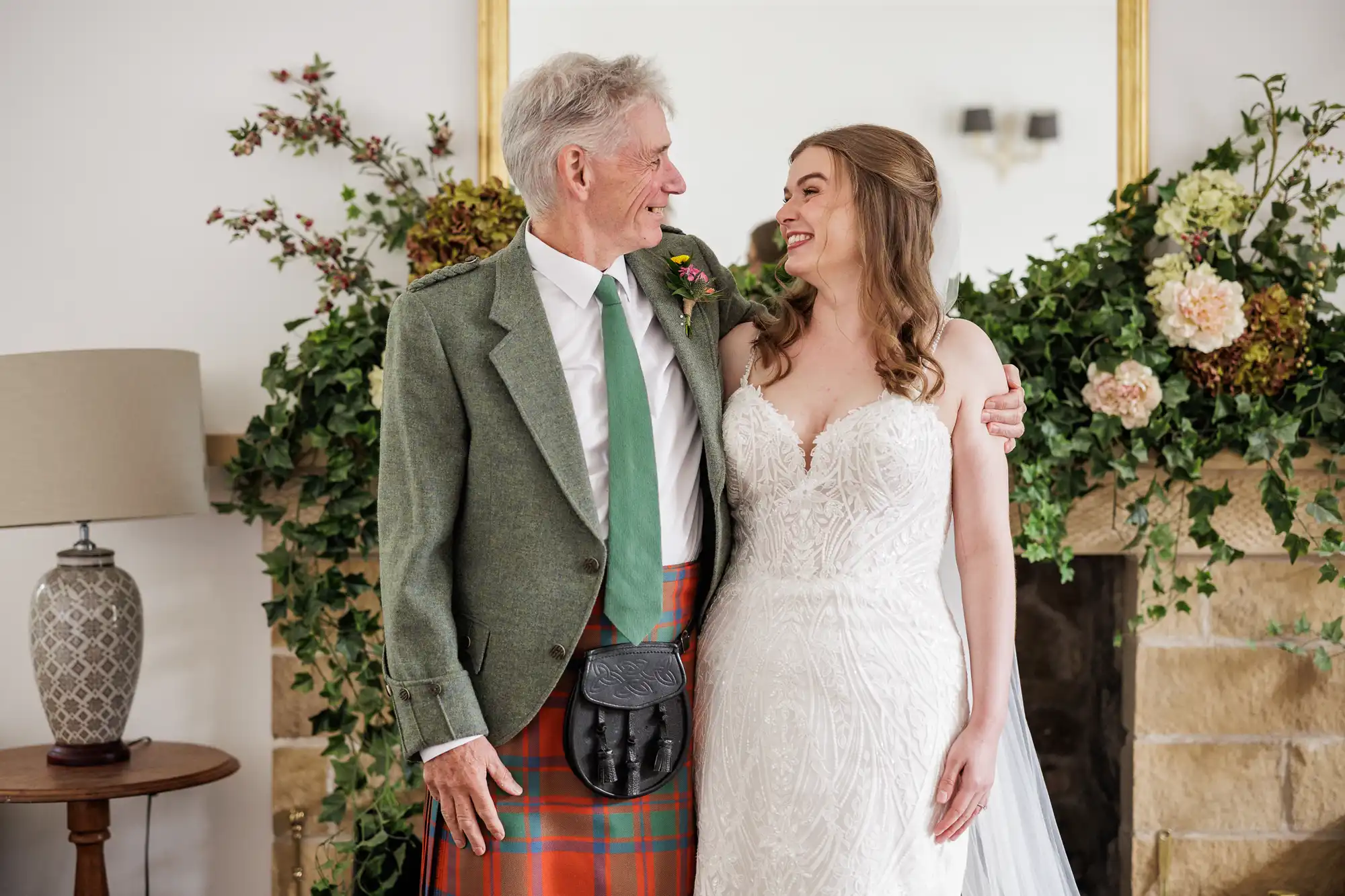 An older man in a green jacket, tartan kilt, and green tie smiles at a woman in a white lace wedding dress. They stand arm in arm in front of a floral-decorated fireplace, looking at each other joyfully.