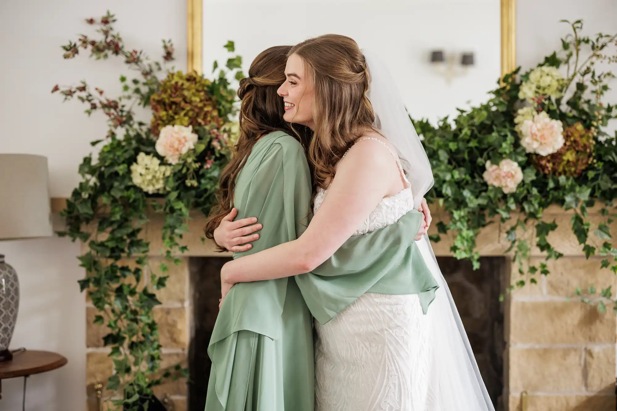 Two women with long brown hair embrace in front of a stone fireplace decorated with lush green ivy and white and pink flowers. One wears a white wedding dress and veil; the other wears a light green dress. Both appear happy, sharing a warm hug.