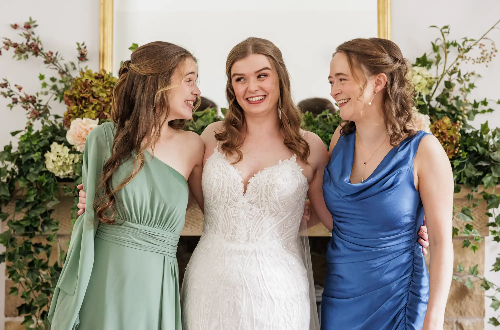 Three women stand arm-in-arm, smiling at one another. The centre woman wears a white lace wedding dress, flanked by women in sage green and royal blue dresses. They are indoors, in front of a mantelpiece decorated with greenery and flowers.