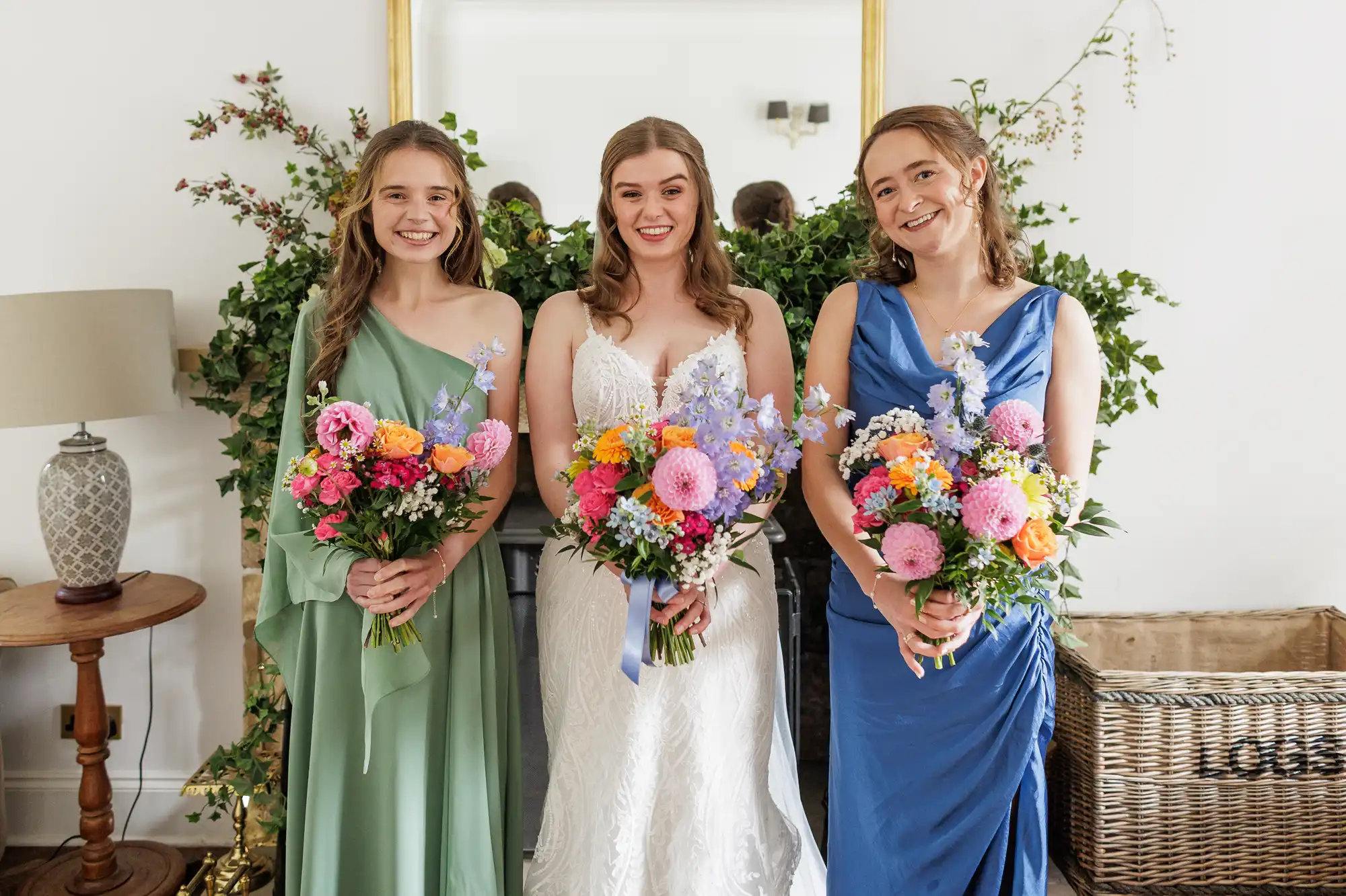 Three women stand indoors, smiling and holding colourful flower bouquets. The woman in the centre wears a white bridal gown, while the women on either side wear green and blue dresses. Behind them is greenery and a mirror, with a lamp and wicker basket nearby.