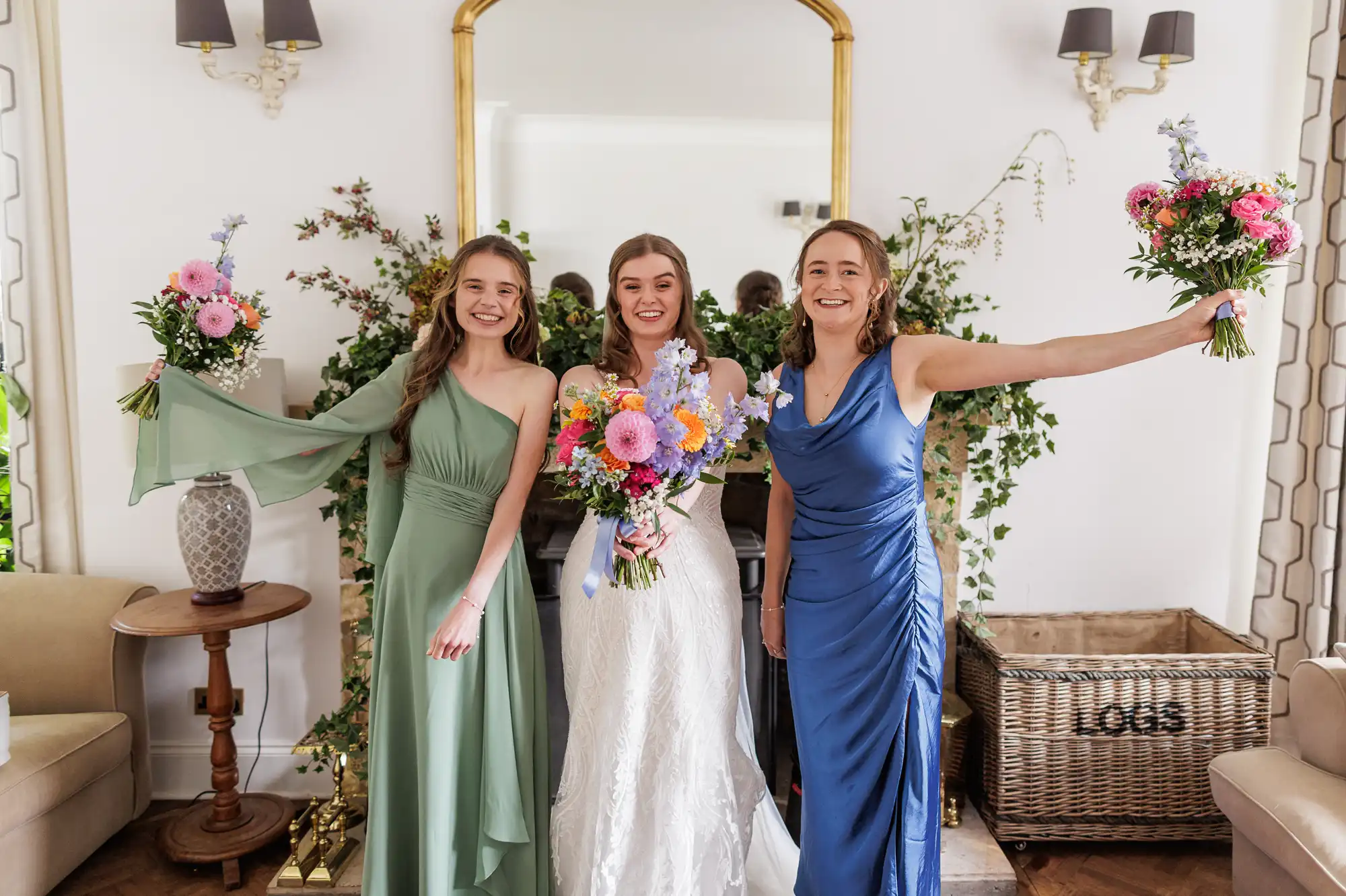 Three women stand smiling indoors. The woman in the middle wears a white wedding dress, holding a bright bouquet. The women beside her wear green and blue dresses and each holds a bouquet. They stand in front of a fireplace with greenery and flowers.
