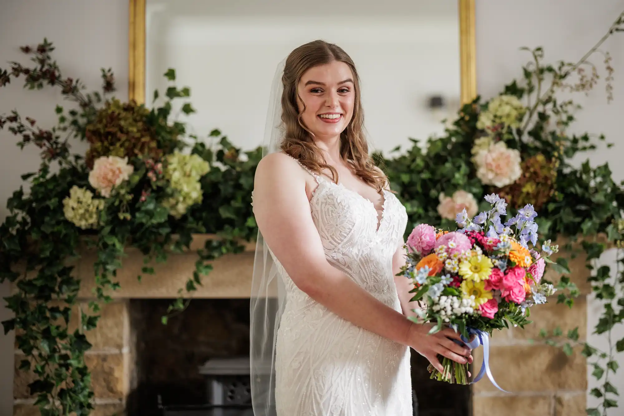 A bride in a white lace gown and veil smiles whilst holding a colourful bouquet of flowers. She stands indoors in front of a fireplace adorned with lush greenery and floral arrangements, with a mirror above the mantelpiece reflecting part of the room.