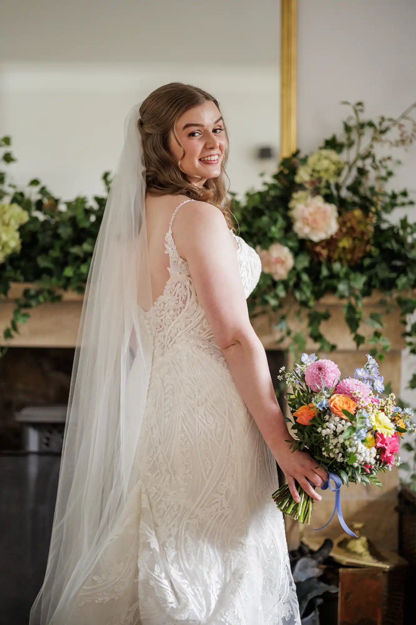A bride in a white lace gown and long veil smiles over her shoulder, holding a colourful bouquet of flowers with a blue ribbon. She stands indoors in front of a fireplace decorated with green foliage and large pink and white flowers.