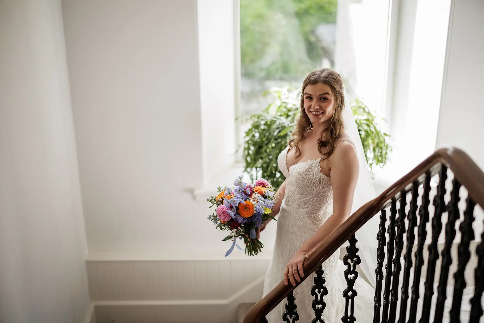 A bride in a white dress stands on a staircase, holding a colourful bouquet of flowers. She smiles, looking slightly to the side, with loose curls framing her face. Behind her, large windows let in natural light and a green potted plant is visible.
