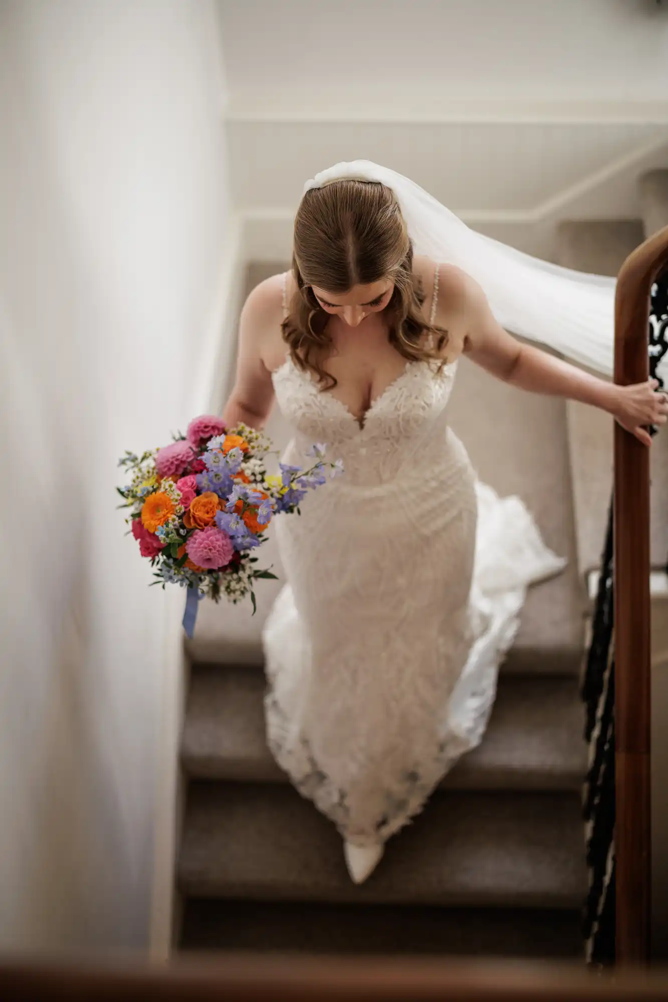A bride in a lace wedding dress and veil descends a carpeted staircase, holding a colourful bouquet of flowers with red, pink, orange, yellow, purple, and blue blooms. She looks down, with one hand on the wooden bannister.