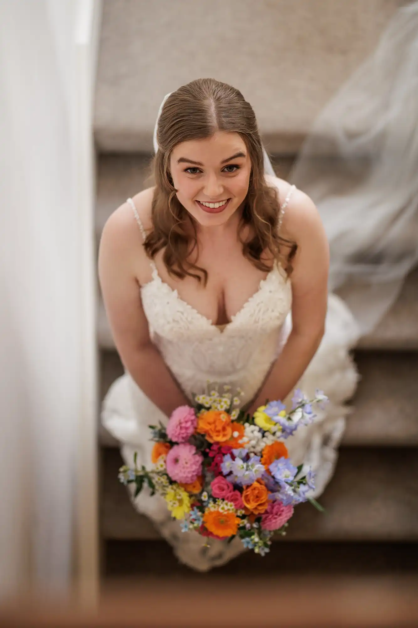 A bride with long brown hair, wearing a white lace wedding dress, sits on carpeted stairs holding a colourful bouquet of flowers. She looks up and smiles, her veil trailing behind her on the stairs. The image is taken from above.