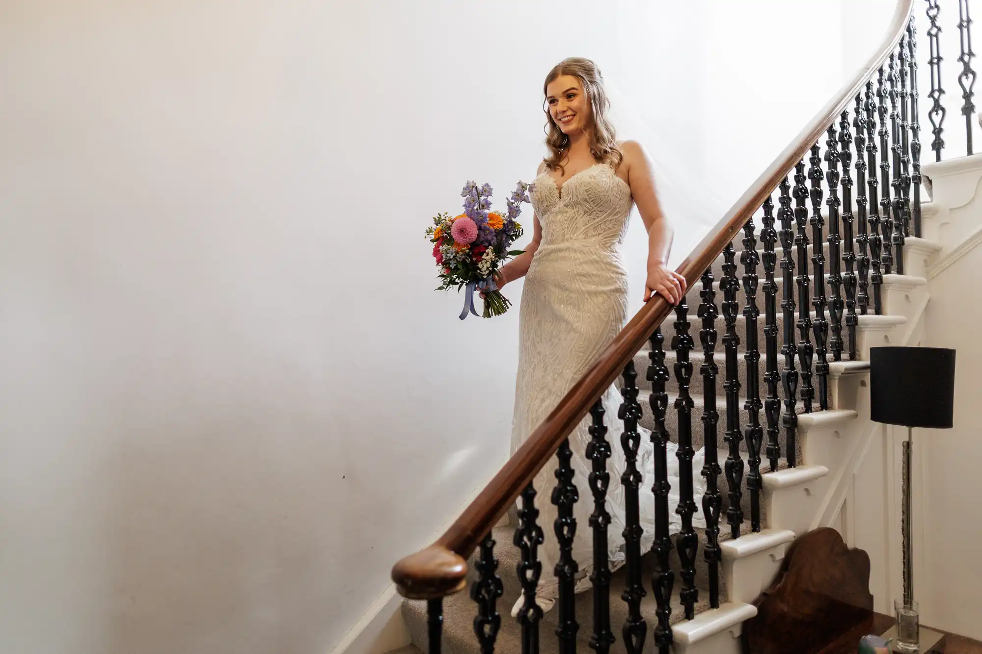 A bride in a white lace wedding dress holds a colourful bouquet whilst descending a curved staircase with dark, ornate railings. She smiles, with loose hair and soft curls, and light streams in, highlighting the simple, elegant setting.