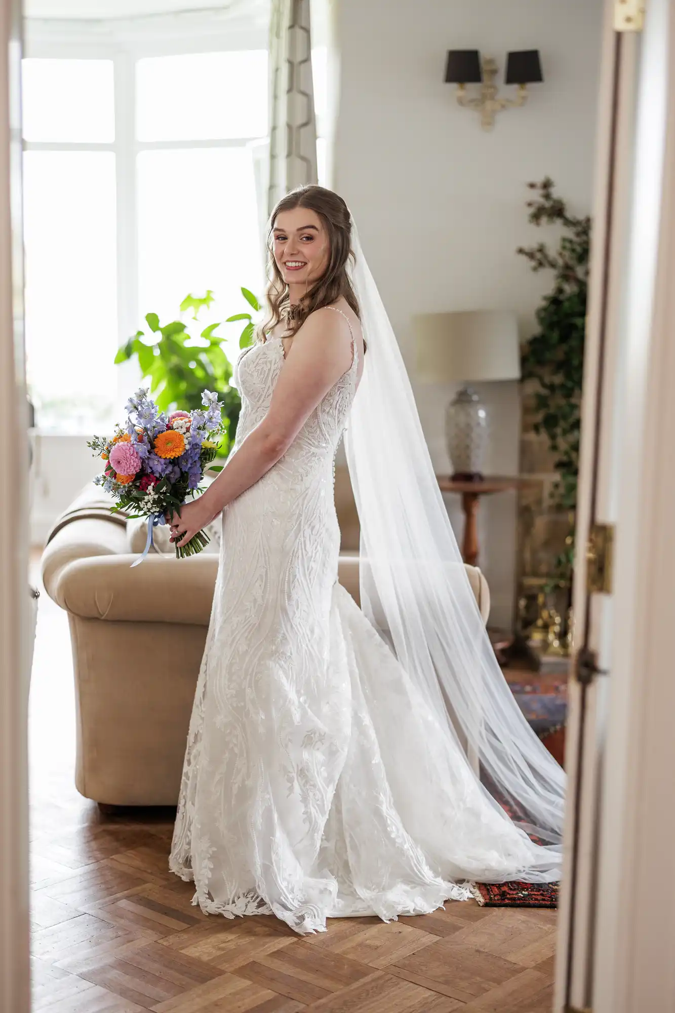 A smiling bride in a fitted, lace white wedding dress stands indoors, holding a colourful bouquet of flowers. Her long veil drapes behind her. She is in a bright room with a beige sofa, large windows, green plants, and wooden flooring.