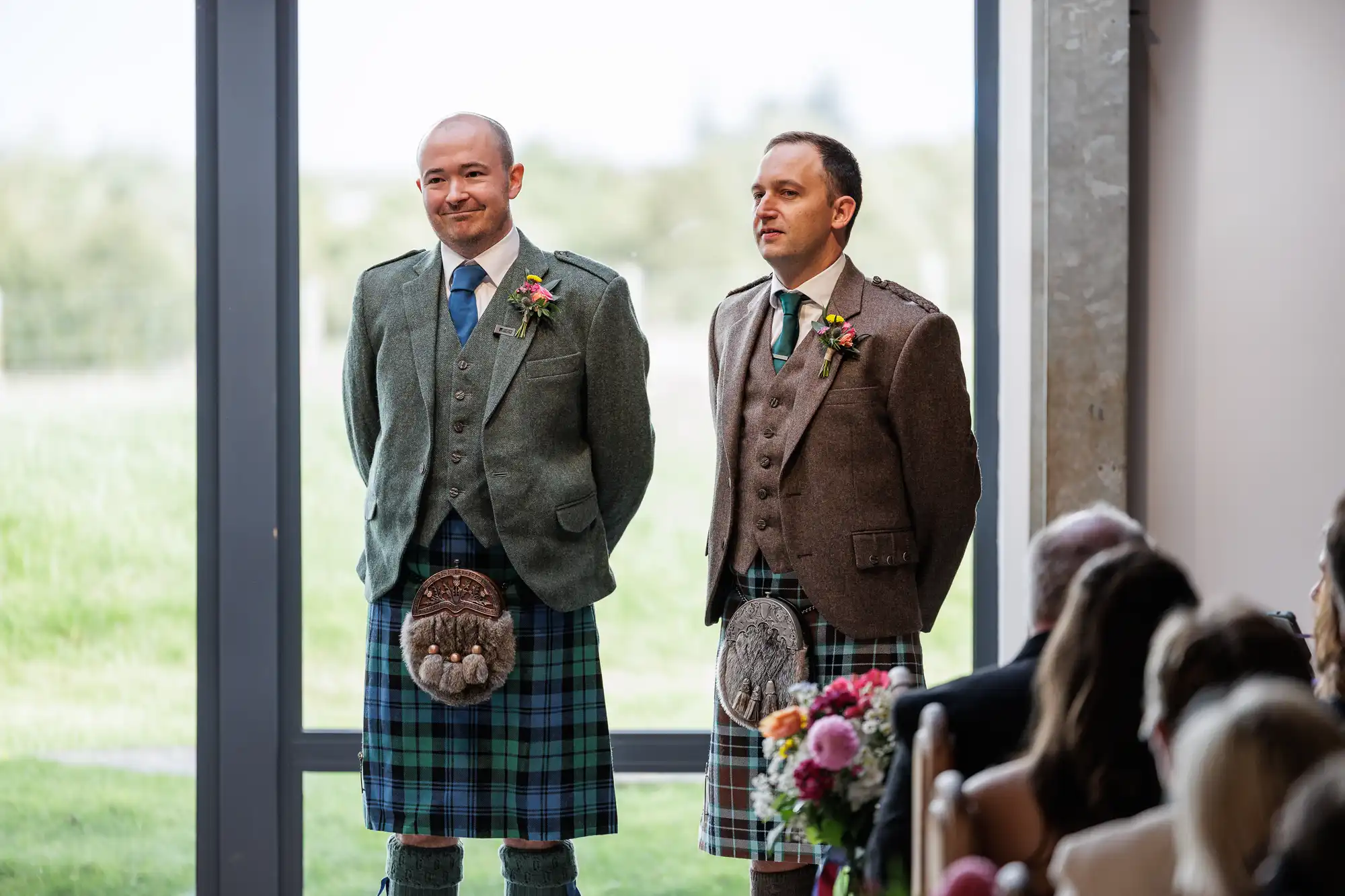 Two men in kilts and tweed jackets stand indoors by a large window. One wears a green jacket and blue tartan kilt, the other a brown jacket and green tartan kilt. Both have sporrans and buttonholes. Seated guests and floral arrangements are visible in the foreground.