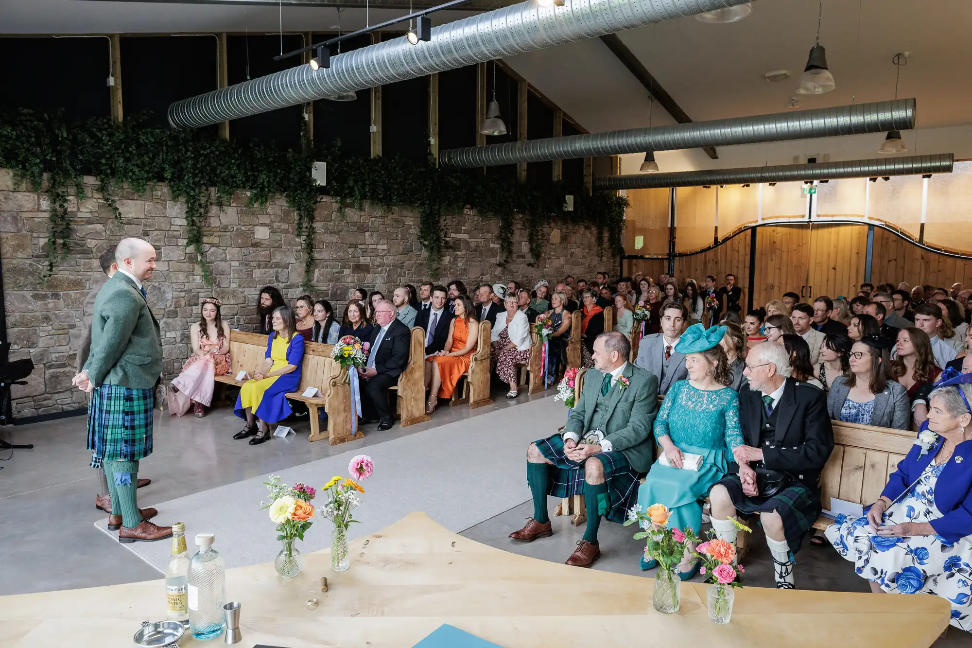 A man in a green jacket and tartan kilt stands addressing a seated wedding audience in a rustic hall with stone walls, wood accents, and greenery. Attendees wear formal attire, some in bright colours. Flower vases decorate a wooden table in the foreground.