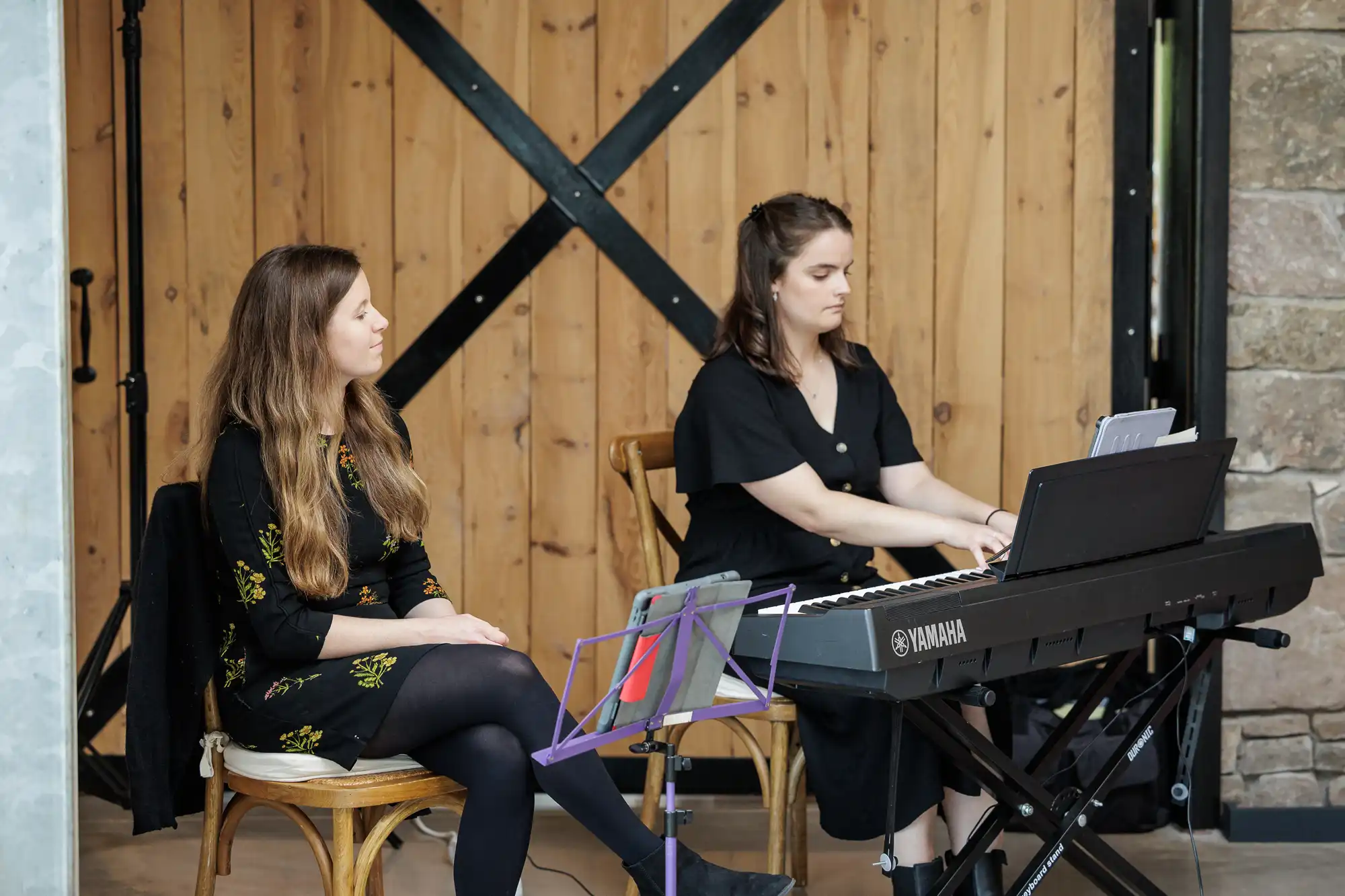 Two women sit indoors; one plays a Yamaha digital piano, focused on sheet music, while the other sits beside her, hands in her lap, looking thoughtful. Both wear black dresses. Behind them are wooden doors with black metal braces and a stone wall.
