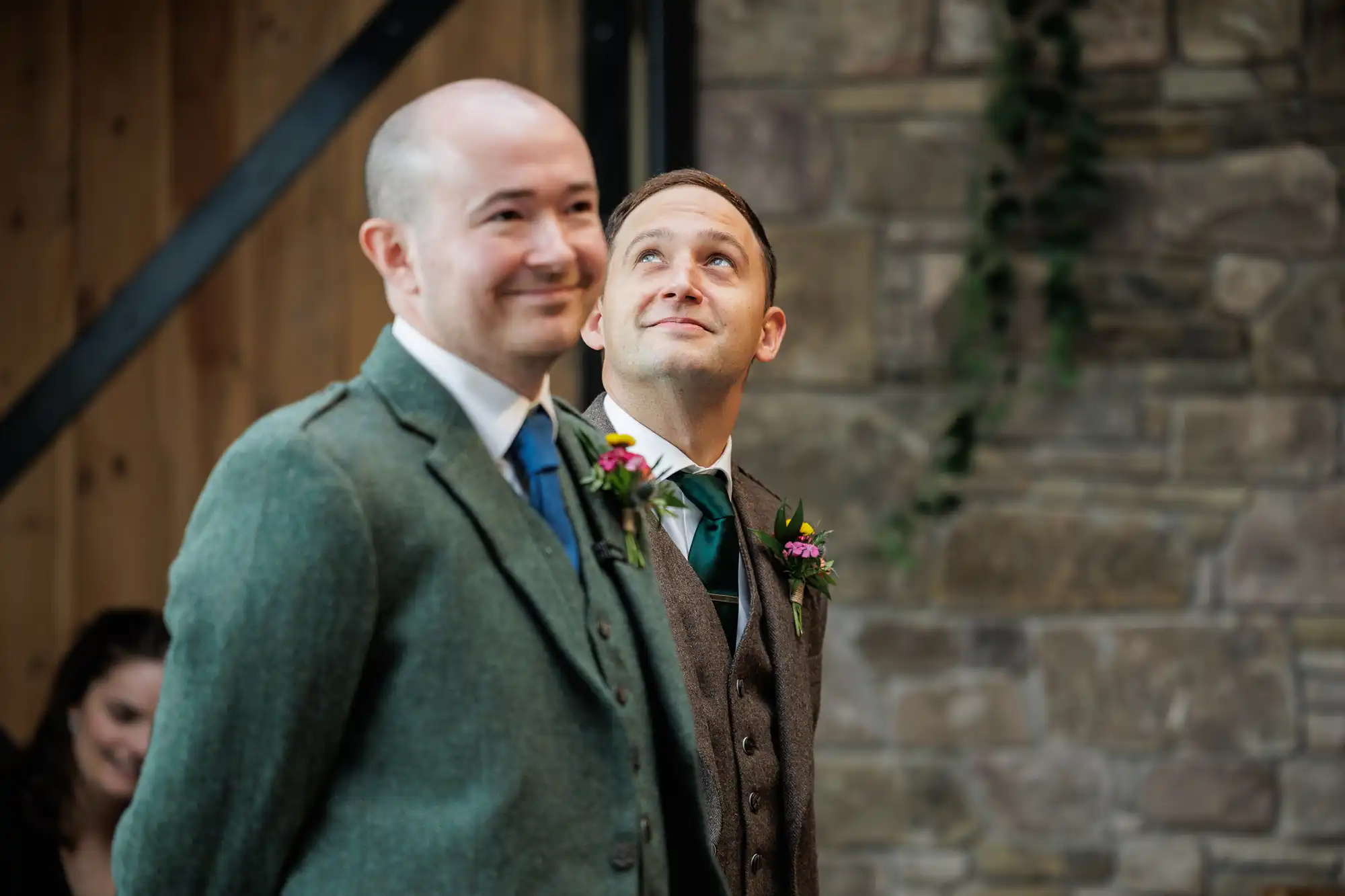 Two men in suits with buttonholes stand indoors, one smiling and the other looking up. The background features a stone wall with climbing greenery and wooden beams. A woman, slightly out of focus, sits in the background, smiling.