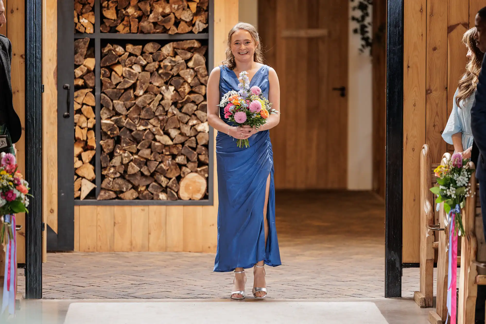A woman in a blue sleeveless dress with a leg slit walks indoors, holding a colourful flower bouquet. She is smiling and wearing silver heels. The background shows stacked firewood, wood-panelled walls, and an open door.