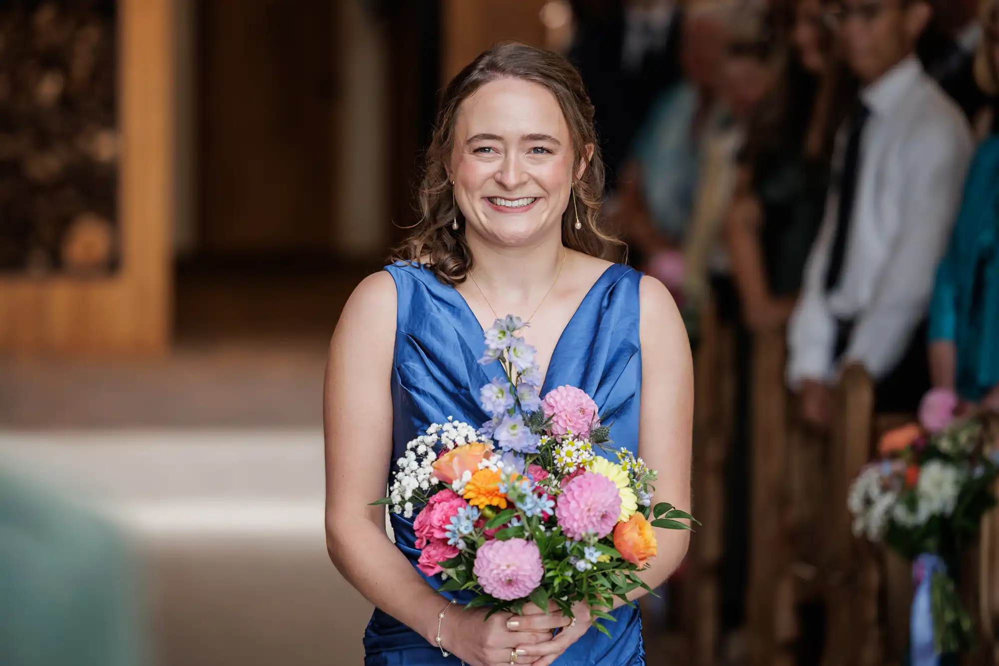 A smiling woman in a royal blue dress holds a colourful bouquet of flowers, including pink, orange, and purple blooms, whilst standing indoors. She is positioned in the aisle, with guests visible and slightly blurred in the background.