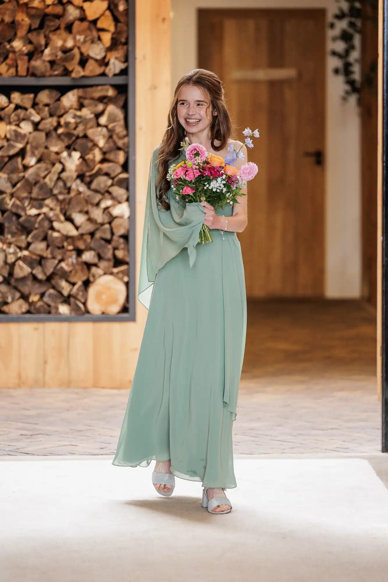 A young woman in a flowing sage green dress and silver sandals walks indoors, smiling and holding a colourful bouquet of flowers. Behind her is a stack of firewood and wooden panelling, with a brown door in the background.