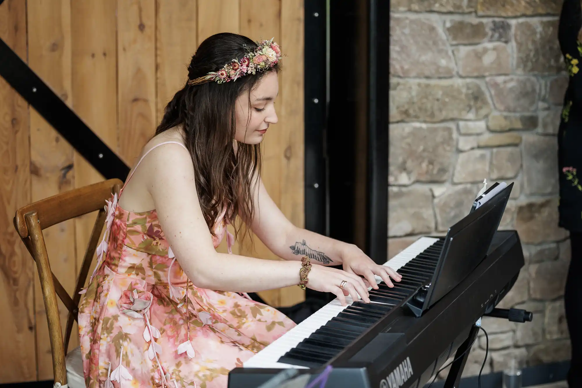 A woman in a floral dress and flower crown sits on a wooden chair, playing a Yamaha keyboard. She has long brown hair and a visible arm tattoo. The background features a wooden door and a stone wall. Sheet music rests on the keyboard stand.