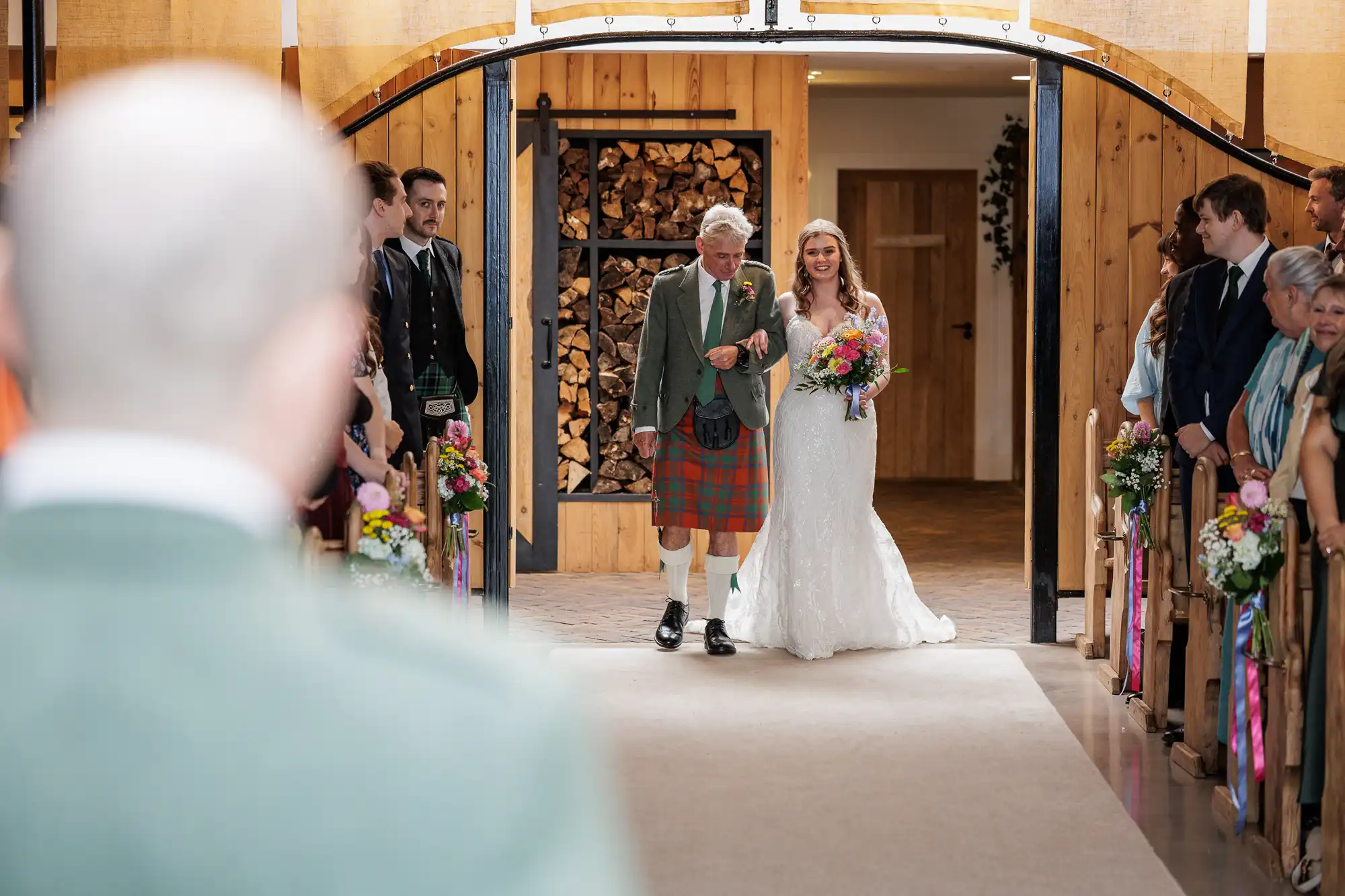A bride in a white dress and veil walks down the aisle with an older man in a kilt, both smiling. Guests sit on either side, some standing, in a warmly lit, wooden hall decorated with flowers. A blurred figure watches in the foreground.