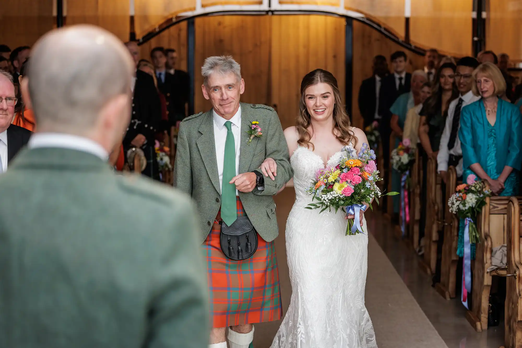 A smiling bride in a white lace gown holds a colourful bouquet and walks down the aisle arm-in-arm with an older man in a green jacket and red tartan kilt. Guests watch from pews adorned with flowers in a warmly lit venue.
