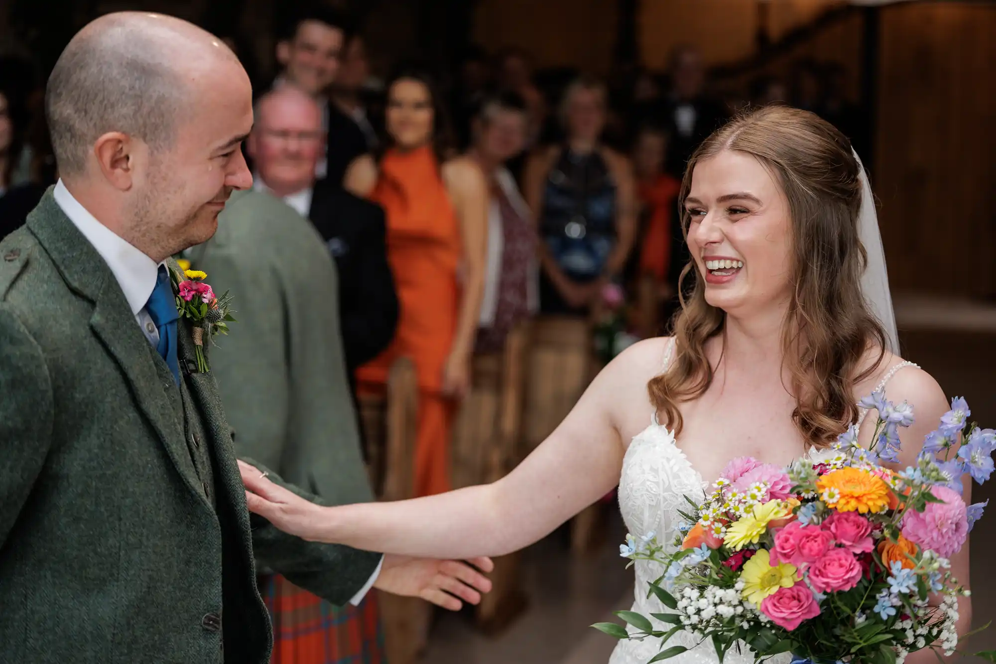 A bride in a white dress, holding a colourful bouquet, smiles joyfully at a groom in a green suit jacket. She touches his chest affectionately. Wedding guests in festive attire are seated and watching in the blurred background.