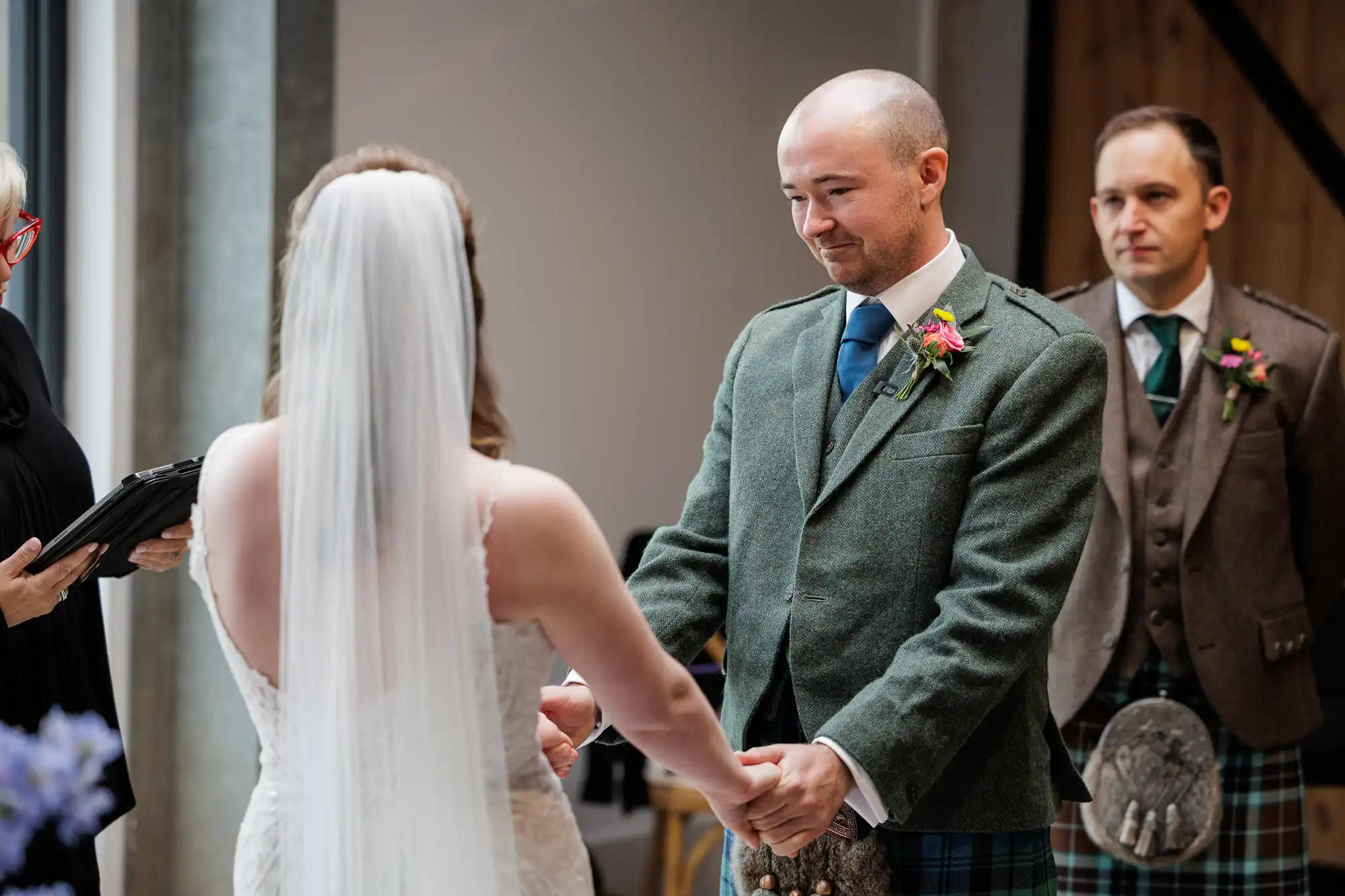 A bride in a white dress and veil holds hands with a groom in a green wool jacket, tartan kilt, and blue tie. Another man in similar attire stands nearby. The setting appears to be indoors, during a wedding ceremony.