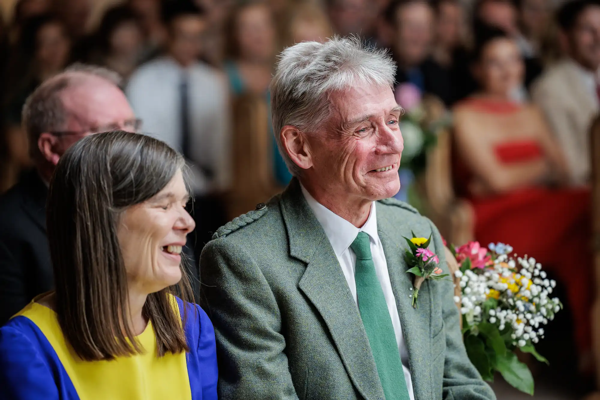 A smiling older couple sit together at an event. The man wears a green suit with a buttonhole and green tie, and the woman has straight brown hair and wears a yellow and blue dress. They are surrounded by people sitting and blurred flowers can be seen behind them.