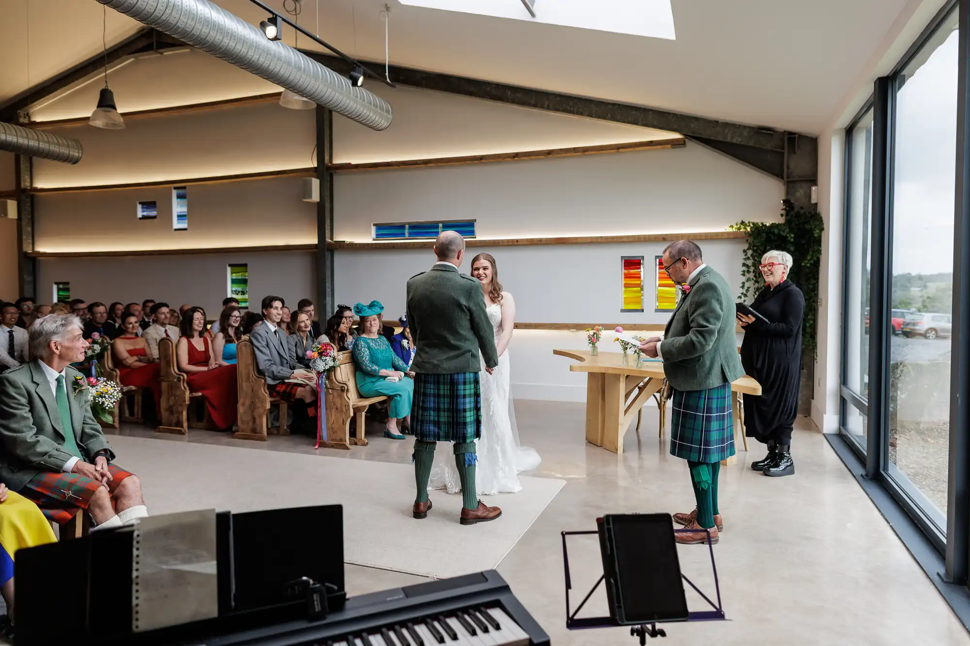 A wedding ceremony in a modern hall with large windows. The bride in a white dress and the groom in a kilt stand facing each other, with guests seated and watching. Two men in kilts and a celebrant stand nearby. Colourful art decorates the walls.