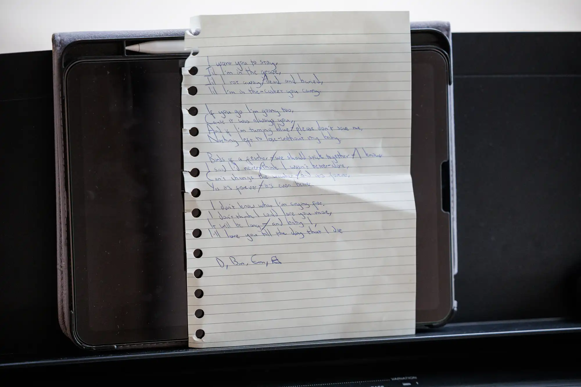 A sheet of lined, hole-punched paper with handwritten song lyrics in blue ink rests on an open black tablet case atop a keyboard. The lyrics are slightly slanted and the page is mildly creased down the centre.