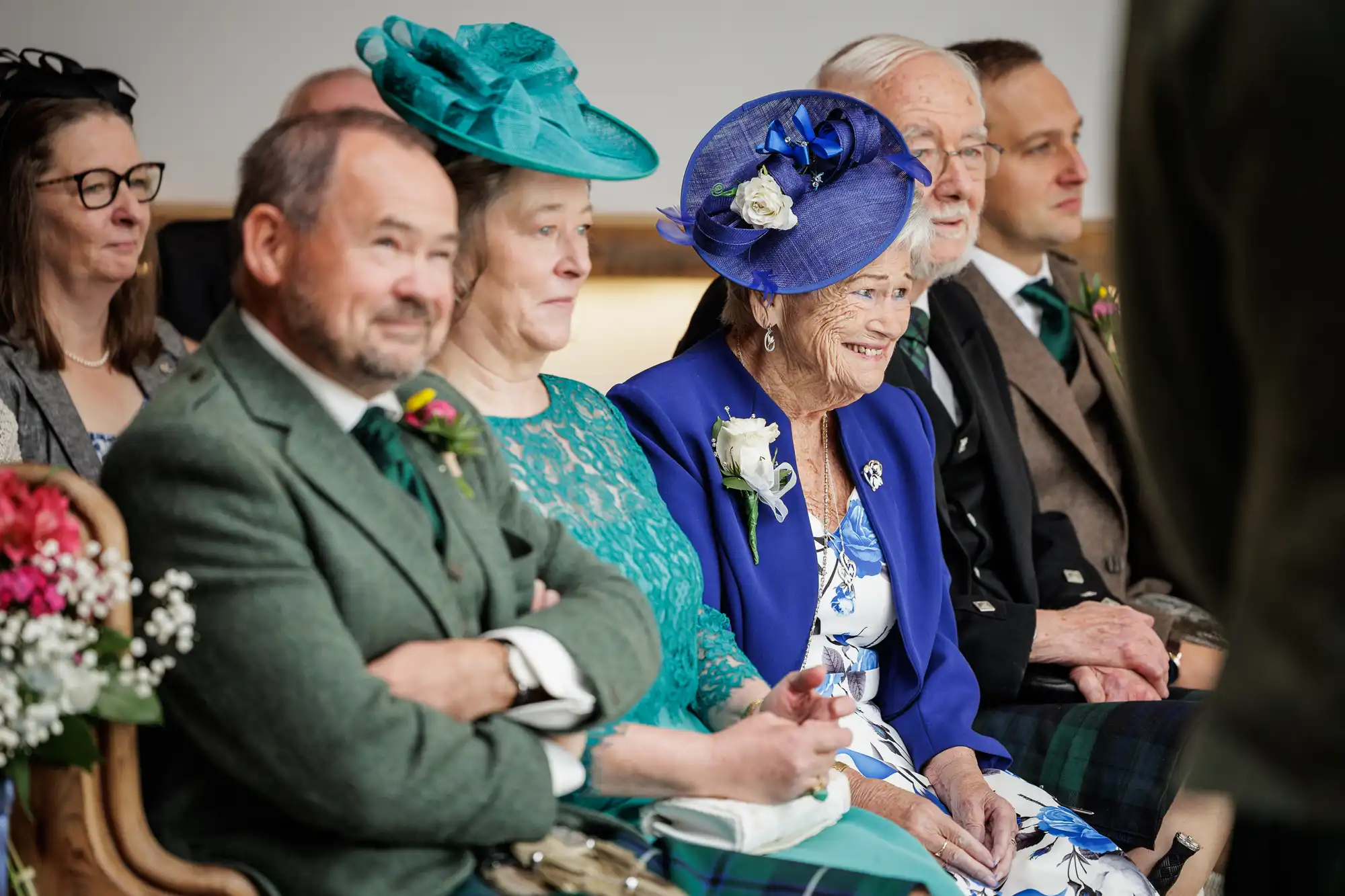 A group of older adults in formal attire sit in a row at an event. Two women wear teal and blue hats; men wear green jackets and tartan kilts. Everyone looks towards the front, smiling and engaged. Flowers and buttonholes decorate their outfits.