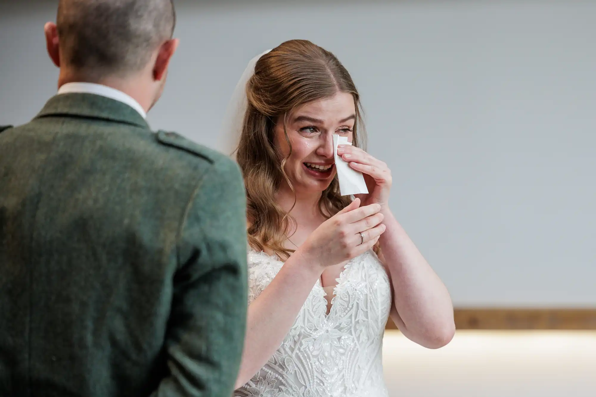 A bride in a white lace wedding dress wipes tears from her eyes with a tissue, smiling emotionally. She has long, wavy hair and a veil. A man in a green suit stands facing her with his back to the camera. The setting is bright and softly lit.