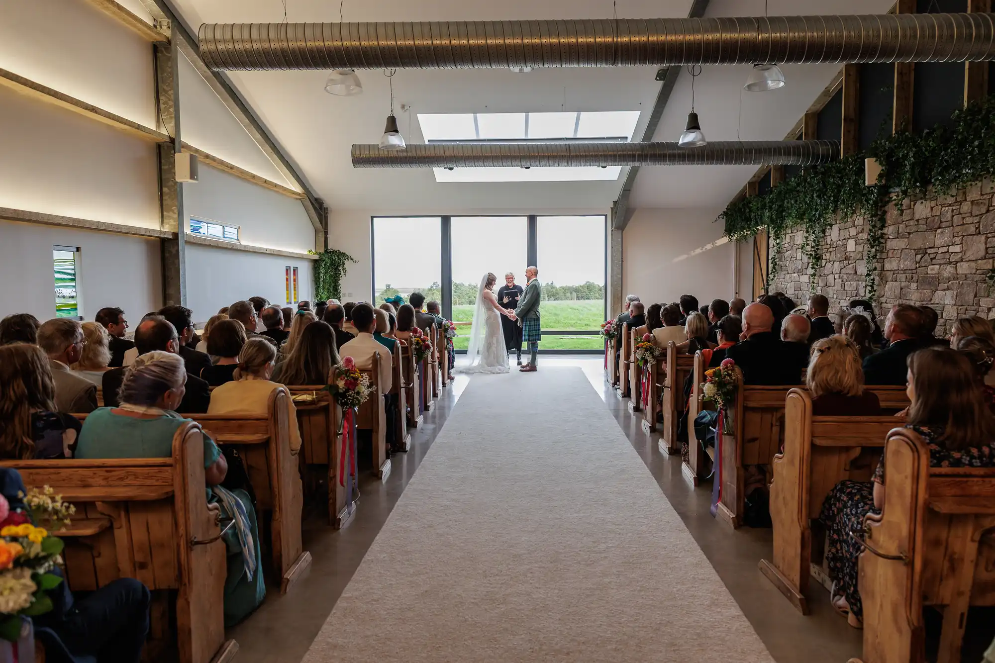 A bride and groom stand at the front of a chapel with large windows, holding hands as an officiant leads their wedding ceremony. Guests are seated in wooden pews on both sides, watching the couple. Greenery decorates the walls, and daylight fills the space.