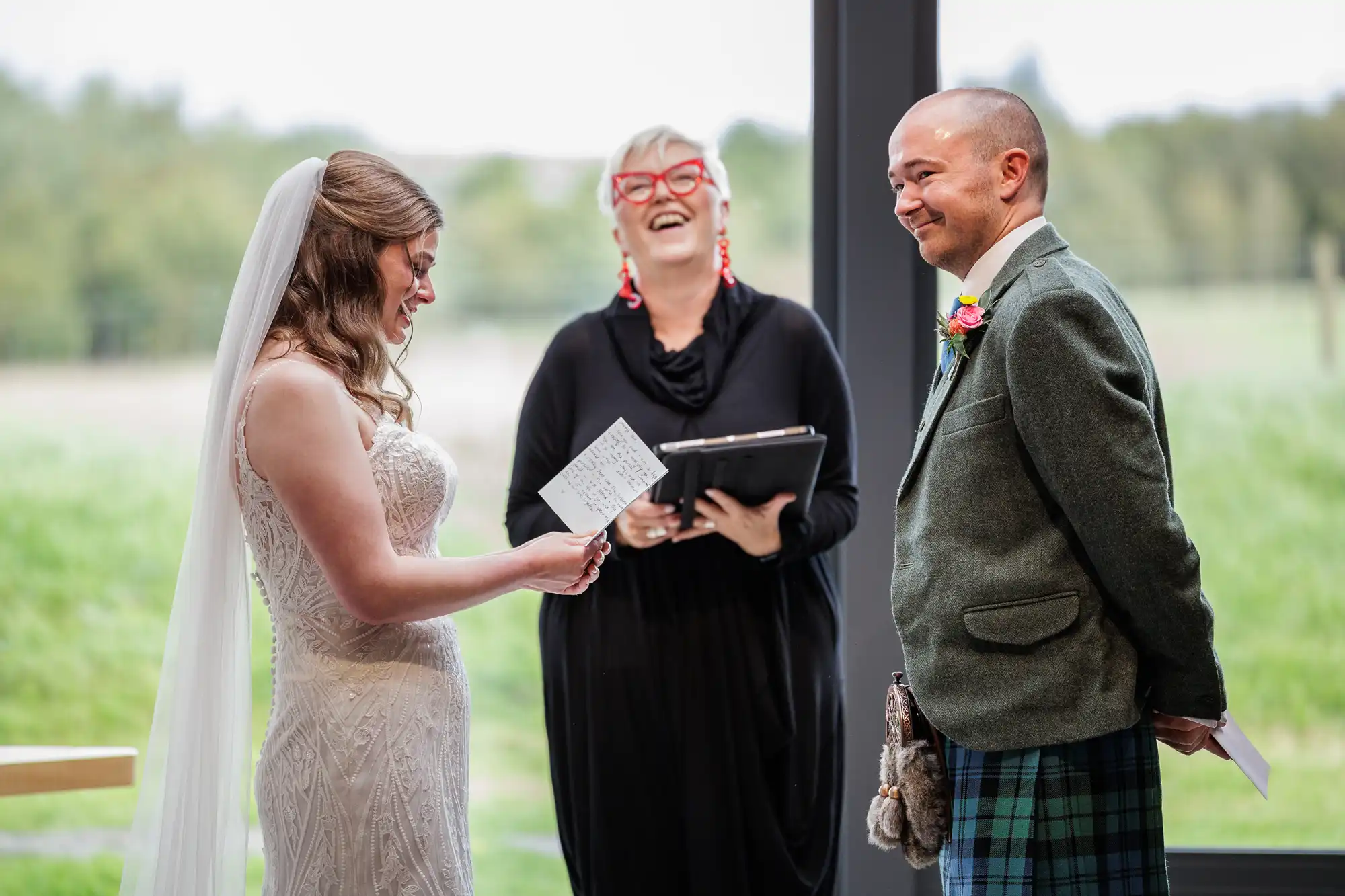 A bride in a white dress and veil reads from a card, smiling at the groom in a kilt and grey jacket. An officiant with short blonde hair, red glasses, and black outfit stands between them, laughing. Large windows show a green, blurred outdoor background.