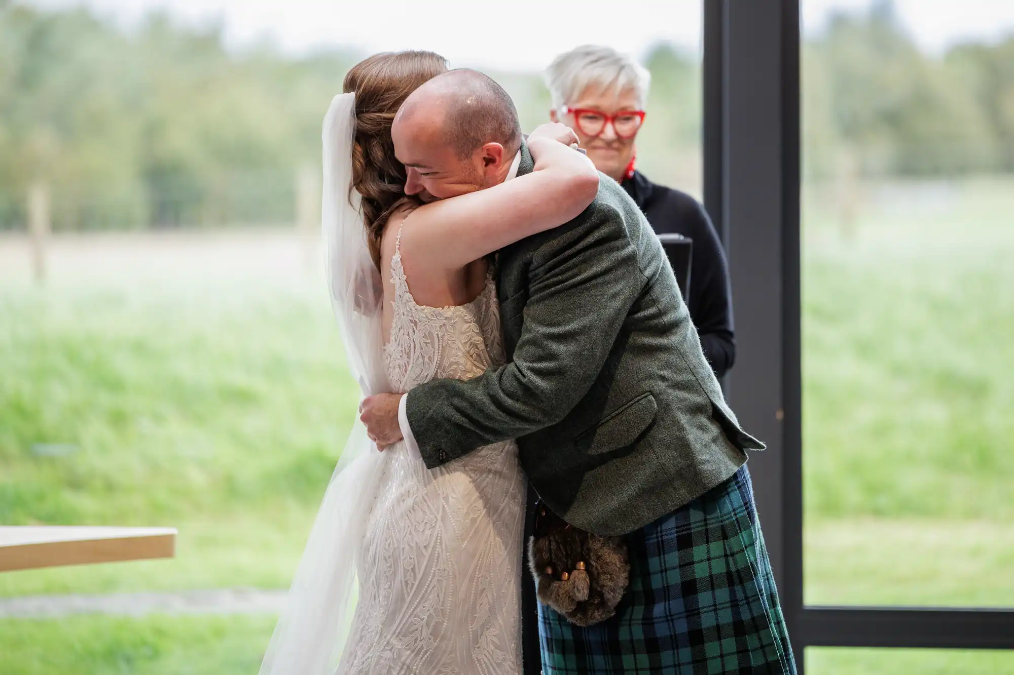 A bride in a white dress and veil hugs a man in a grey jacket and green tartan kilt. Both appear emotional. A woman with short grey hair and red glasses stands in the background, smiling. Outside, green grass and trees are visible through large windows.