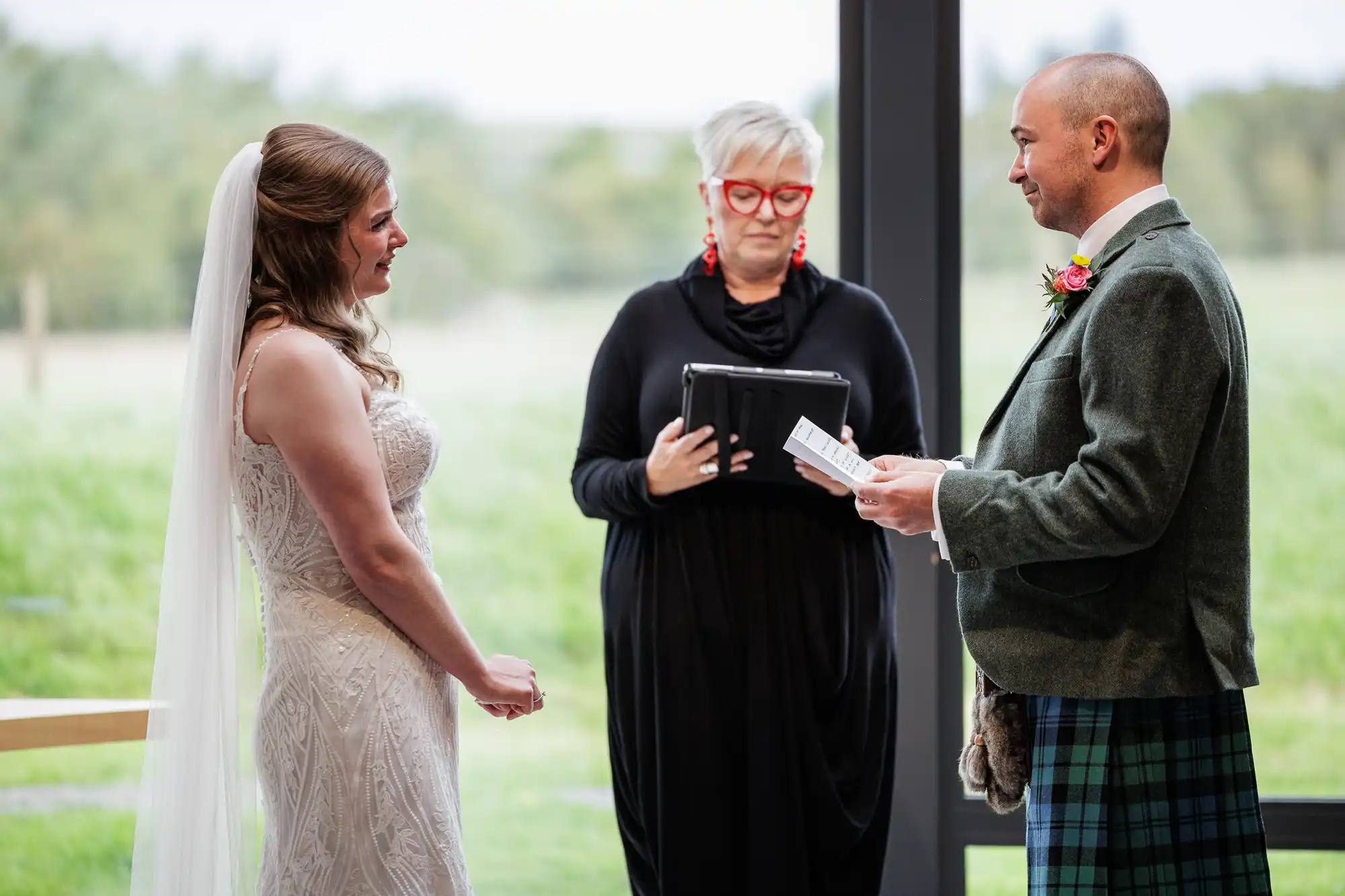 A bride in a lace dress and veil smiles at a groom in a green jacket and tartan kilt, who is reading vows. An officiant in a black outfit with red glasses and earrings stands between them, holding a tablet. Green landscape is visible through large windows behind them.
