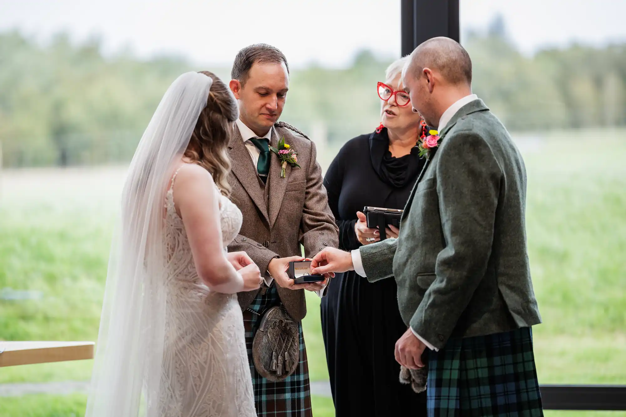 A bride in a lace dress and veil faces a groom in a tweed jacket and kilt. An officiant in black and a man in a green jacket and kilt hold rings. Large windows behind show greenery outside. All appear focused on the exchange.