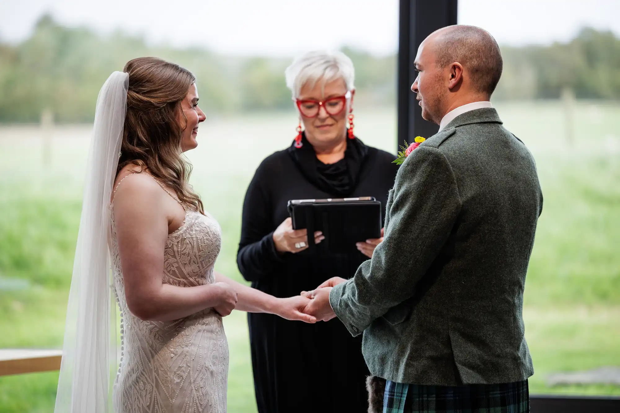 A bride in a strapless white dress and veil holds hands with a groom in a green jacket and kilt. They face each other, smiling, while an officiant with short white hair, glasses, and red earrings reads from a tablet. A grassy landscape is visible outside the window.