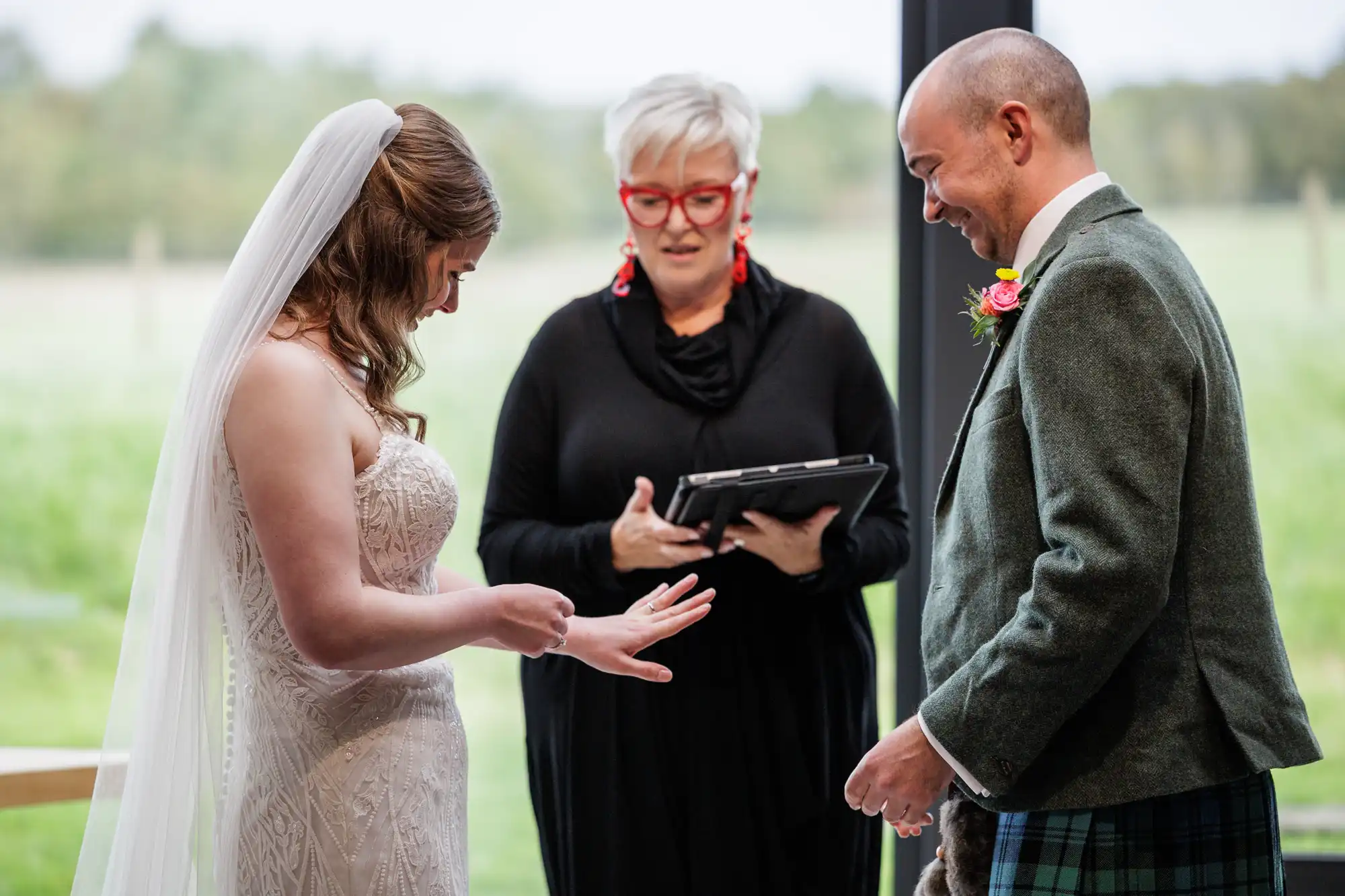 A bride in a white dress and veil admires her ring while smiling at a groom in a green suit and kilt. An officiant in black, holding a tablet, stands between them. Large windows reveal a grassy landscape outside.