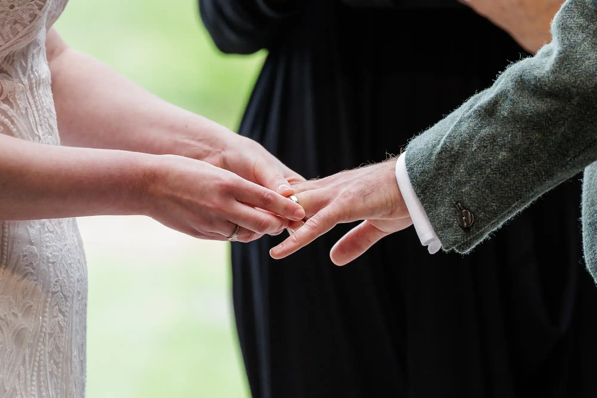 Close-up of a bride placing a ring on her partner’s finger during a wedding ceremony. The bride wears a white lace dress, while the partner wears a green jacket. A third person, blurred in black, stands in the background.