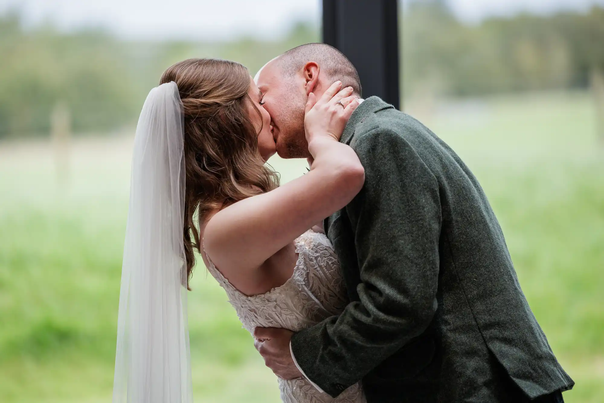 A bride in a white lace dress and veil kisses a groom in a green suit jacket. They embrace closely indoors, the groom holding the bride’s waist and the bride’s arms around his neck. Green fields and trees are visible through large windows behind them.