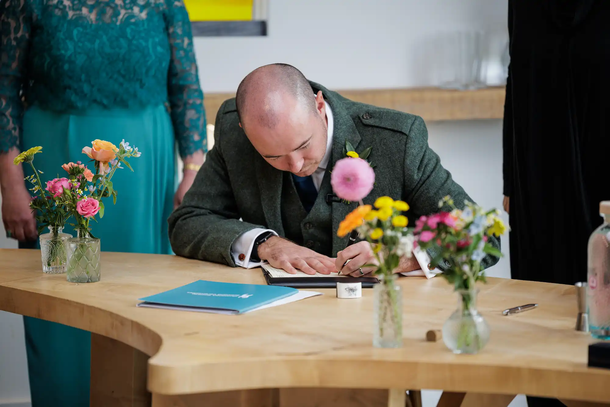 A man in a green suit signs a document at a wooden table decorated with colourful flowers in glass jars. Two women stand nearby, one wearing a teal lace dress. Papers, a pen, and a glass bottle are also on the table. The setting appears formal.
