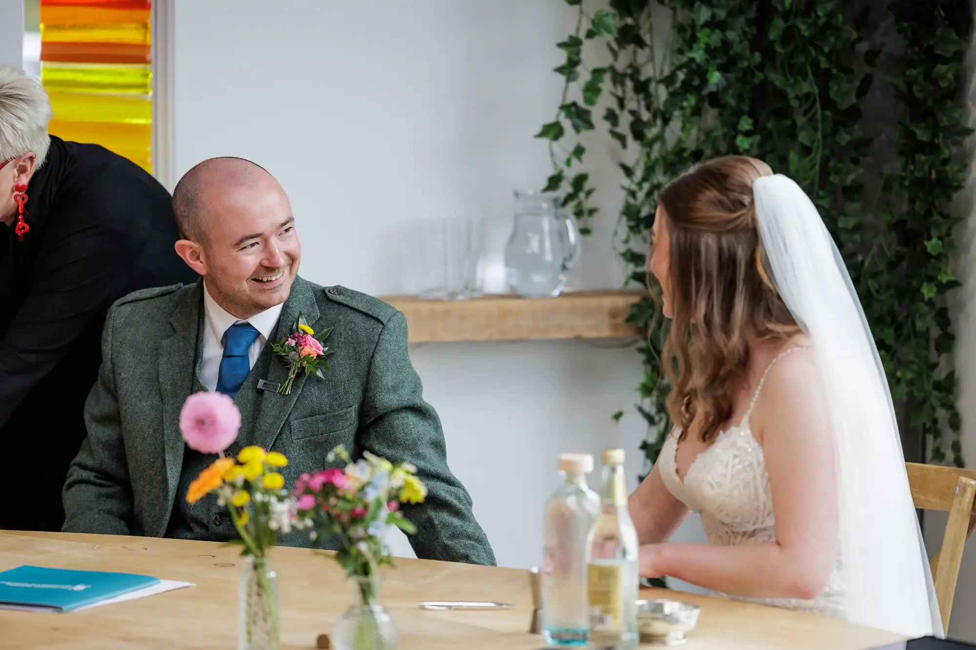 A man in a green suit and blue tie smiles at a woman in a white wedding dress and veil, sitting at a wooden table with flowers, water bottles, and glasses. Greenery hangs in the background, and a colourful artwork is partially visible on the wall.