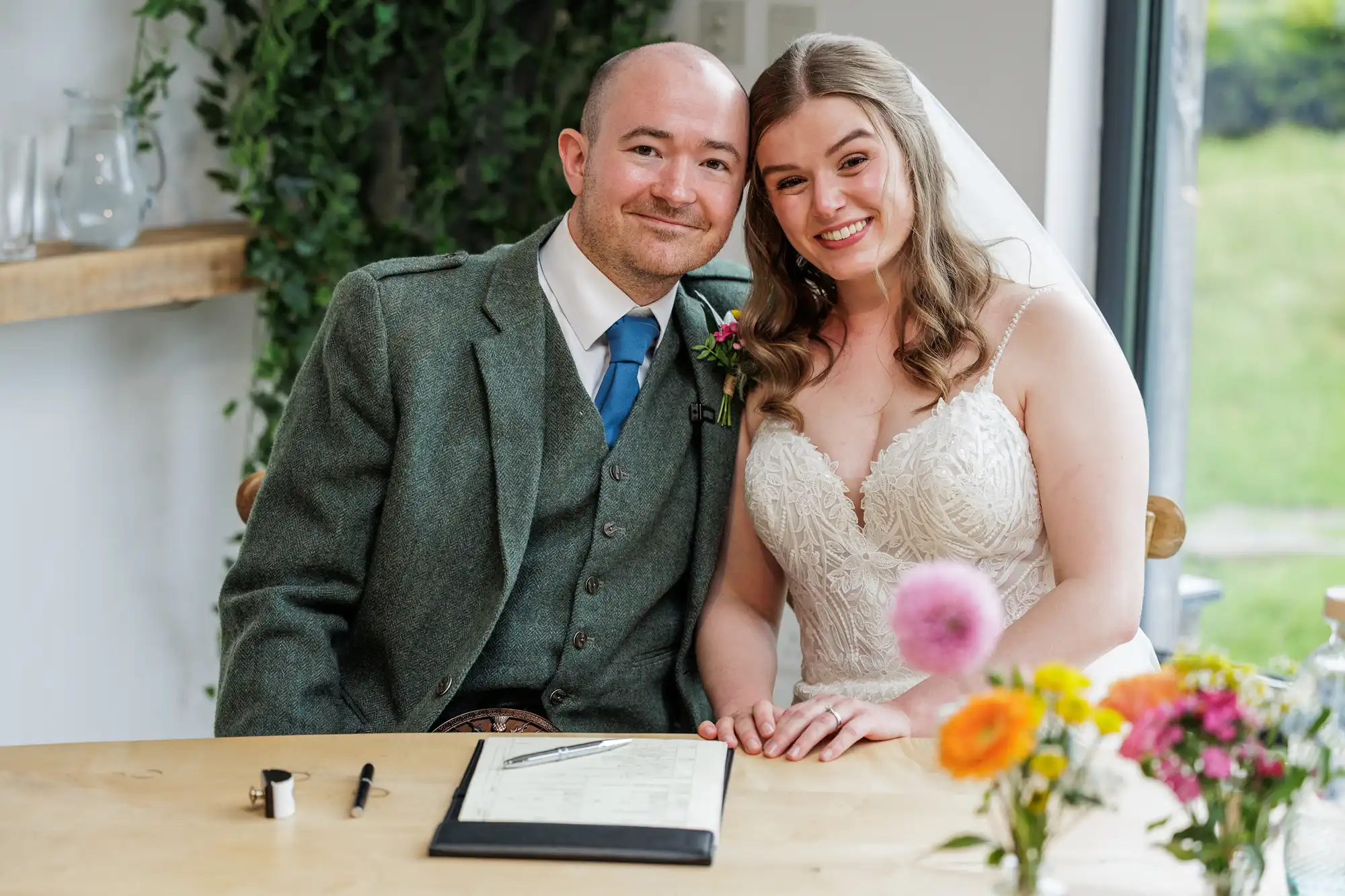 A smiling bride in a white lace gown and a groom in a green suit with a blue tie sit close together at a table, holding hands in front of a signed document. Colourful flowers are in the foreground; greenery and windows are in the background.