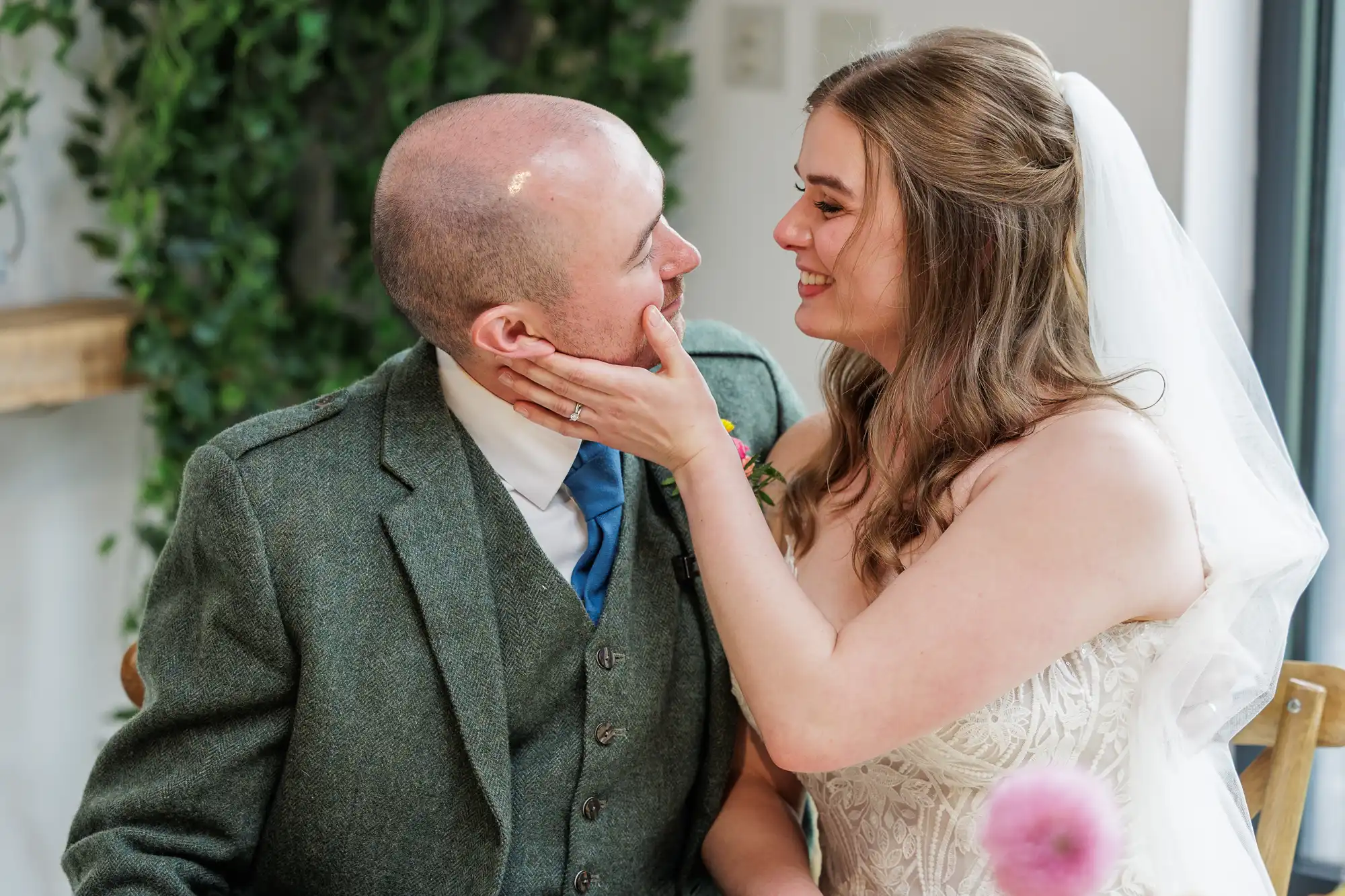 A bride in a white dress and veil smiles lovingly as she gently touches the face of a groom in a green suit. They gaze into each other’s eyes, seated indoors near greenery, sharing a joyful, intimate moment on their wedding day.