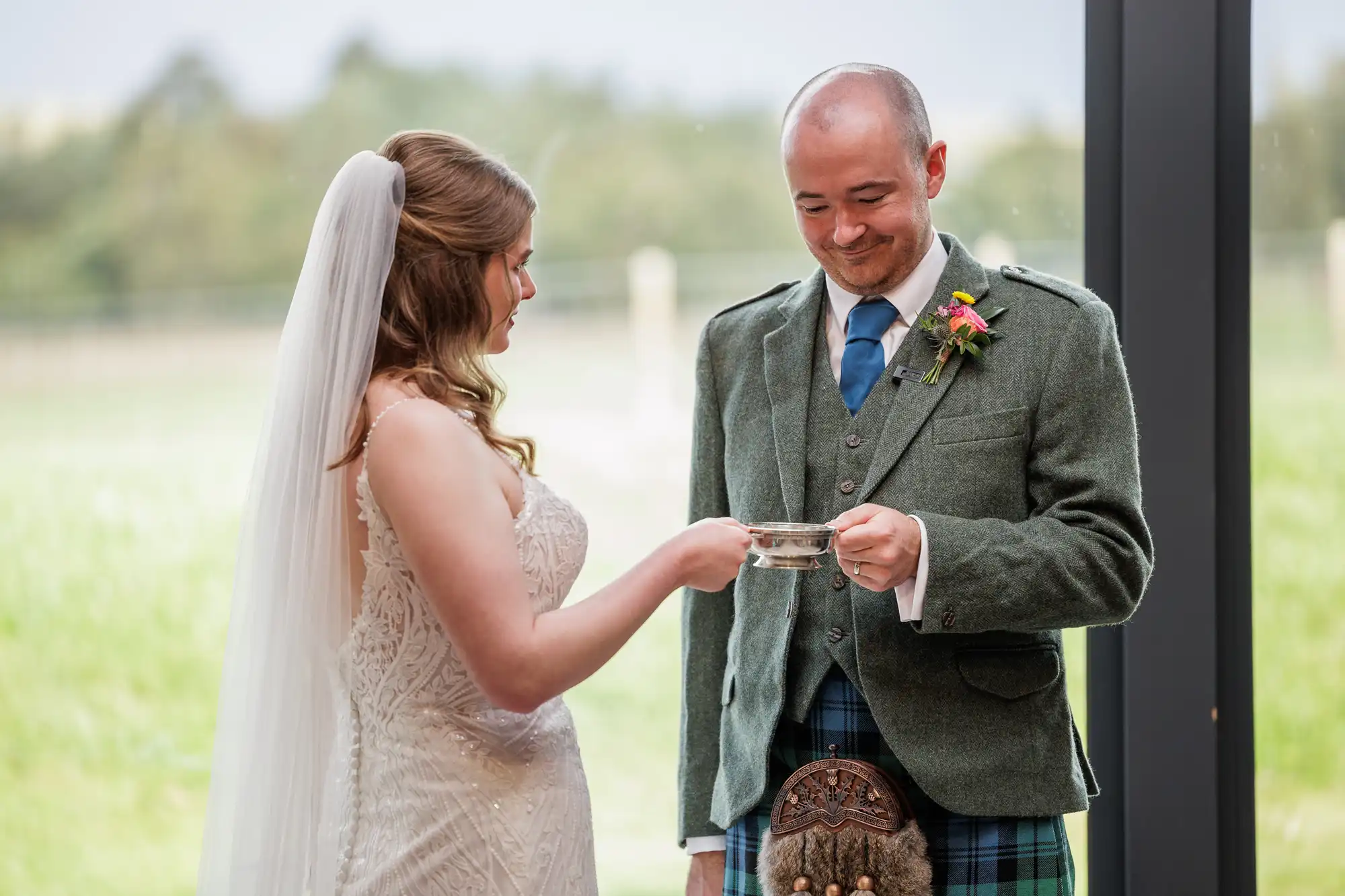 A bride in a white dress and veil and a groom in a green jacket, blue tie, and tartan kilt share a moment by a large window; the bride hands a small cup to the smiling groom, with greenery visible outside.