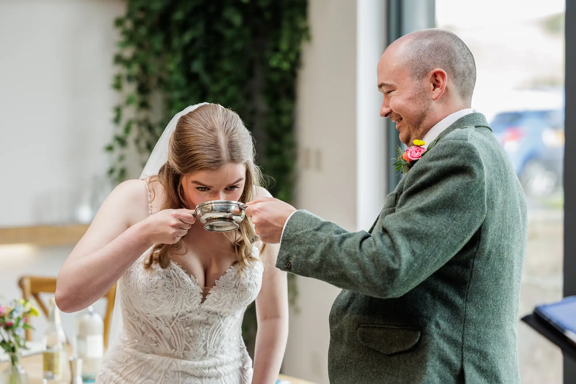 A bride in a white lace dress and veil drinks from a silver cup held by a smiling groom in a green suit jacket with a colourful buttonhole. They stand indoors near a window with greenery visible in the background.