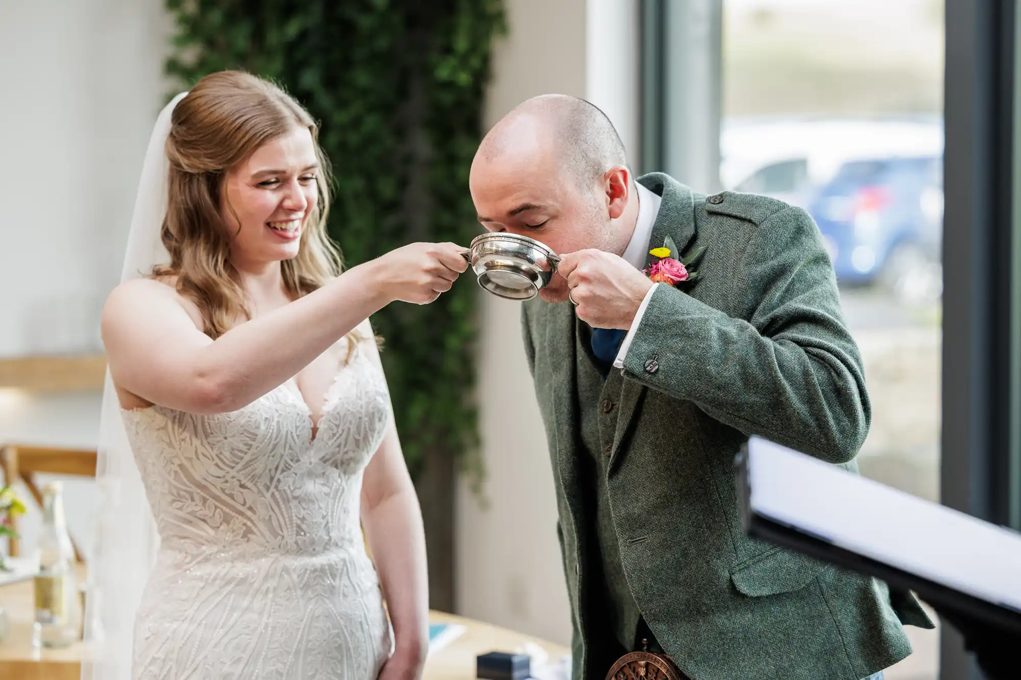A smiling bride in a white dress holds up a silver cup for a groom in a green suit to drink from. They appear happy and are participating in a wedding ritual indoors, with bright natural light coming through large windows behind them.