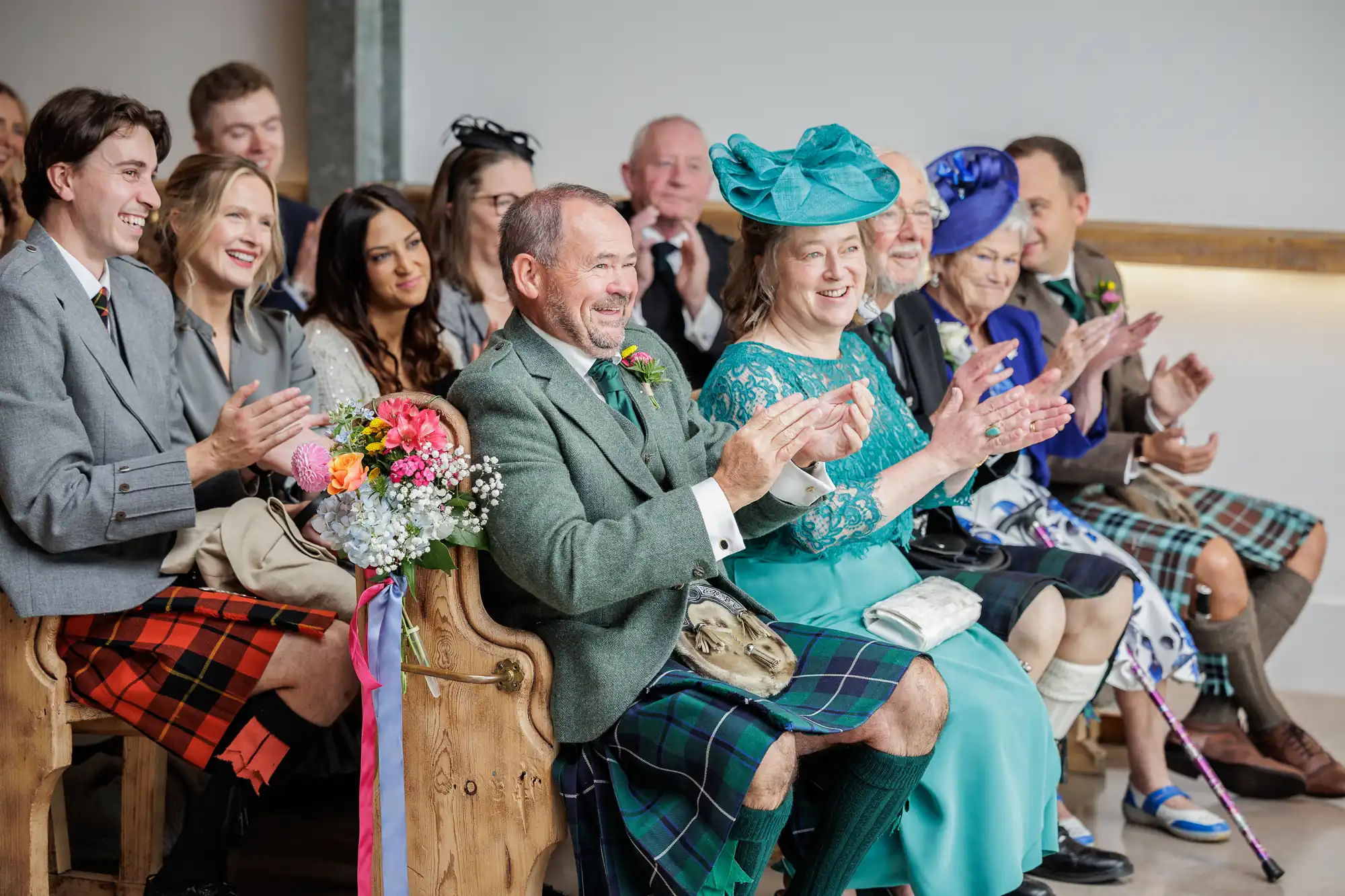 A group of wedding guests in Scottish attire, including kilts and colourful hats, sit on wooden benches, smiling and clapping. The mood is joyful and festive, with floral bouquets and diverse people in formal dress celebrating indoors.