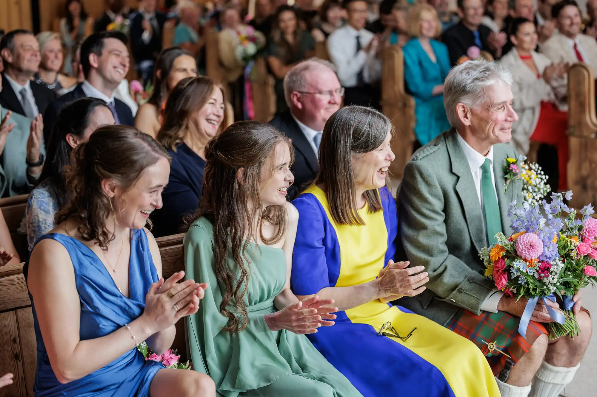 A joyful group of people in colourful formal attire sit on wooden benches, smiling and clapping. A man in a green jacket and tartan kilt holds a bouquet, while women in green, blue, and yellow dresses sit beside him. The audience in the background also appears happy.
