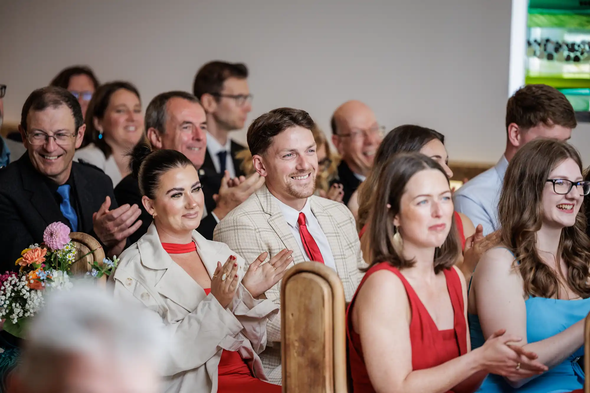 A group of well-dressed people sit in wooden pews, smiling and clapping. The focus is on a man in a light checked suit and red tie, with a woman in red beside him. The mood is joyful, suggesting they are attending a celebratory event, possibly a wedding.