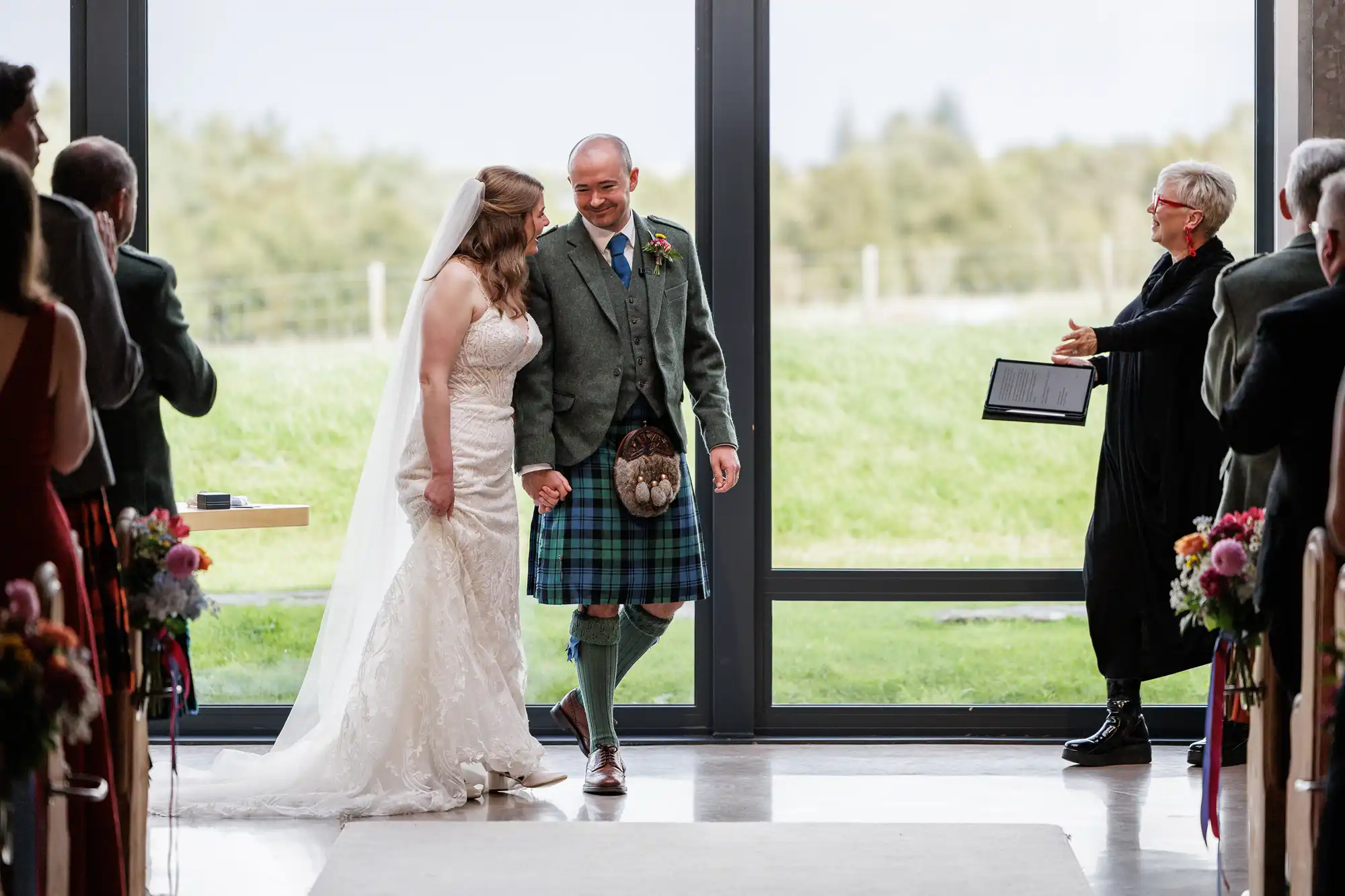 A bride in a white lace gown and veil walks arm-in-arm with a groom wearing a green jacket, blue tartan kilt, and sporran. They smile at each other, heading down the aisle while guests watch and an officiant claps in front of a large window.